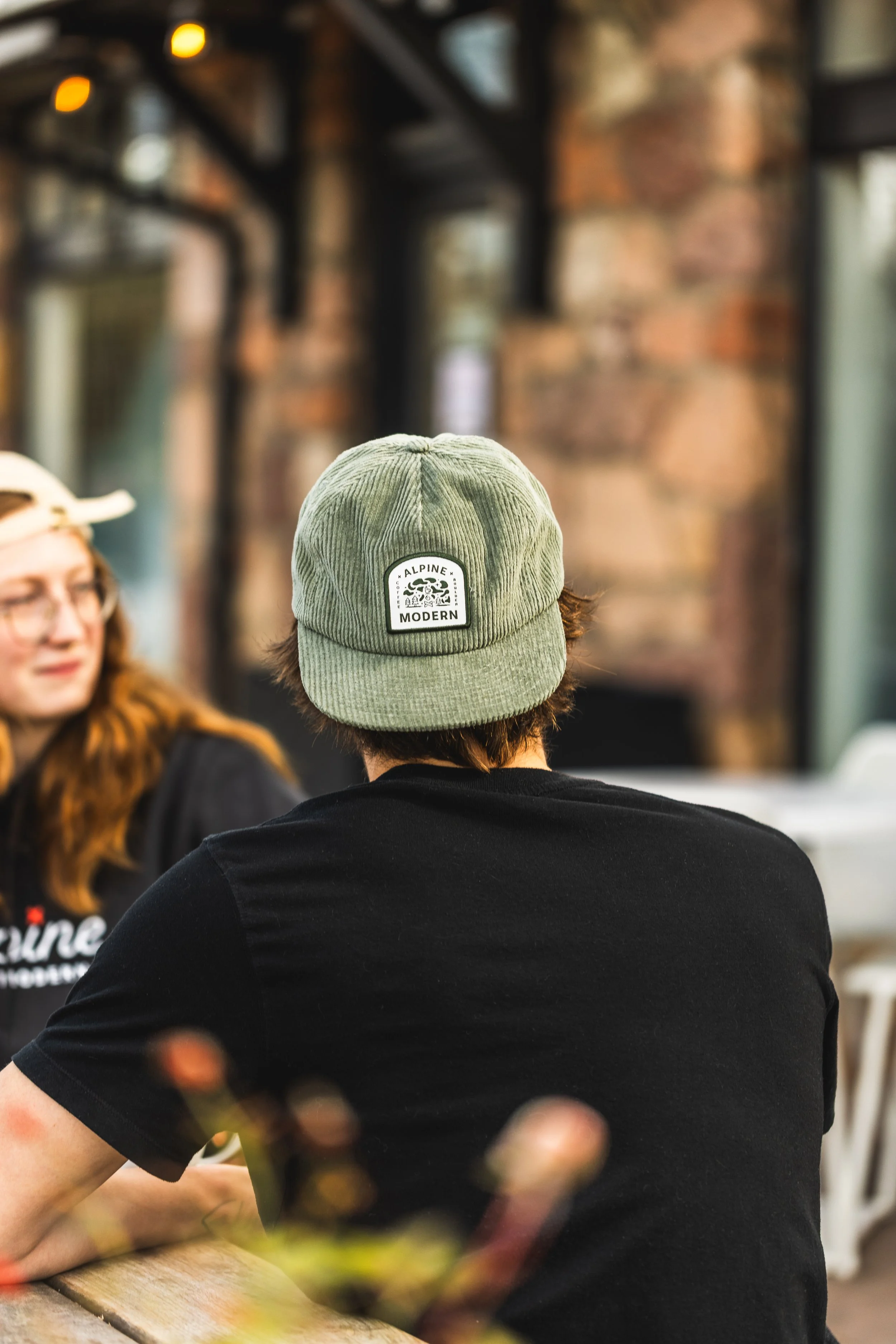 A person wearing a green corduroy baseball cap with a patch that reads "Alpine Modern" seen from behind, sitting at a table in a cafe with brick walls and other people.