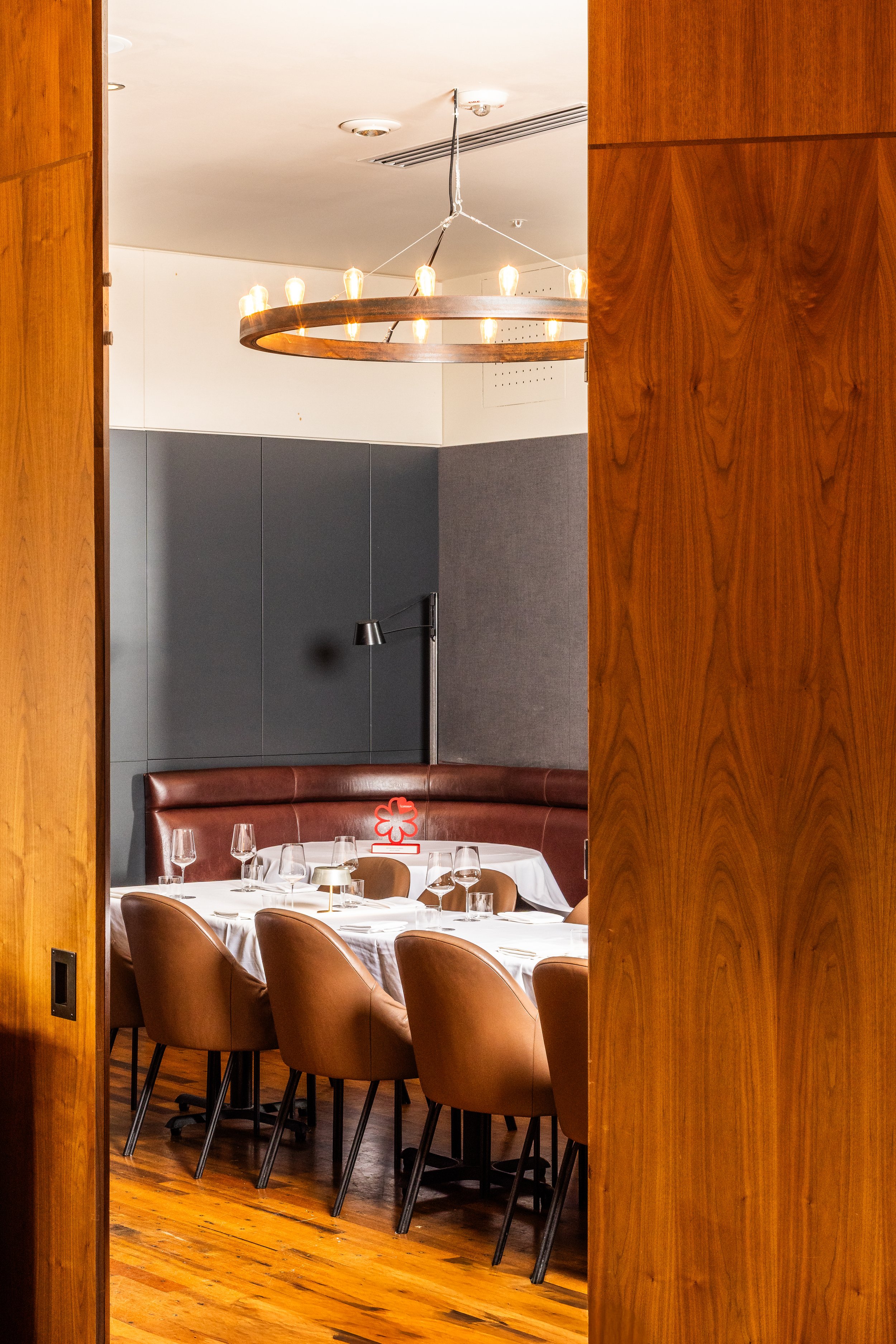 A dining room viewed through an open wooden sliding door, featuring a round table set with wine glasses, a white tablecloth, and brown leather chairs, with a dark gray wall and a modern floor lamp in the background.