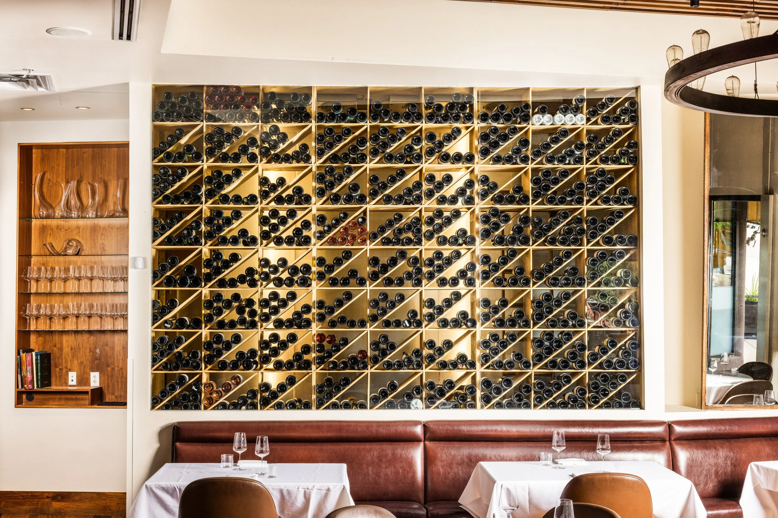Interior of a restaurant with wine bottles stored in a large wooden wine rack covering most of the wall. Tables with white tablecloths and empty wine glasses are in the foreground.