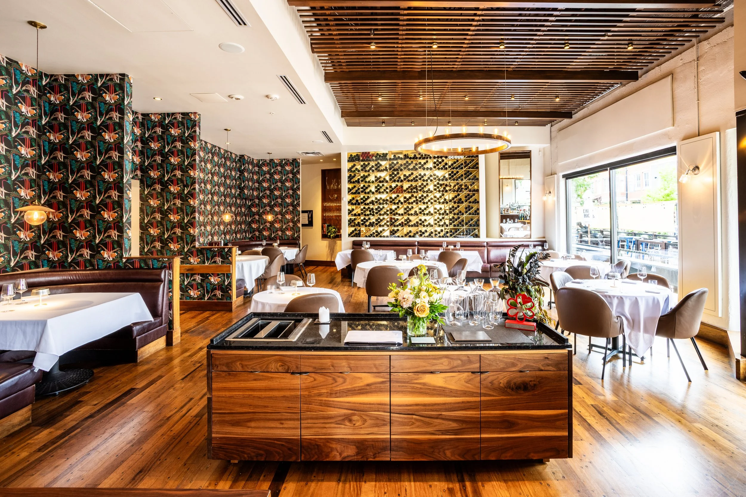 Elegant restaurant interior with wooden floors, white tablecloths, and modern lighting, featuring a wine display on the wall and floral centerpieces on the tables.