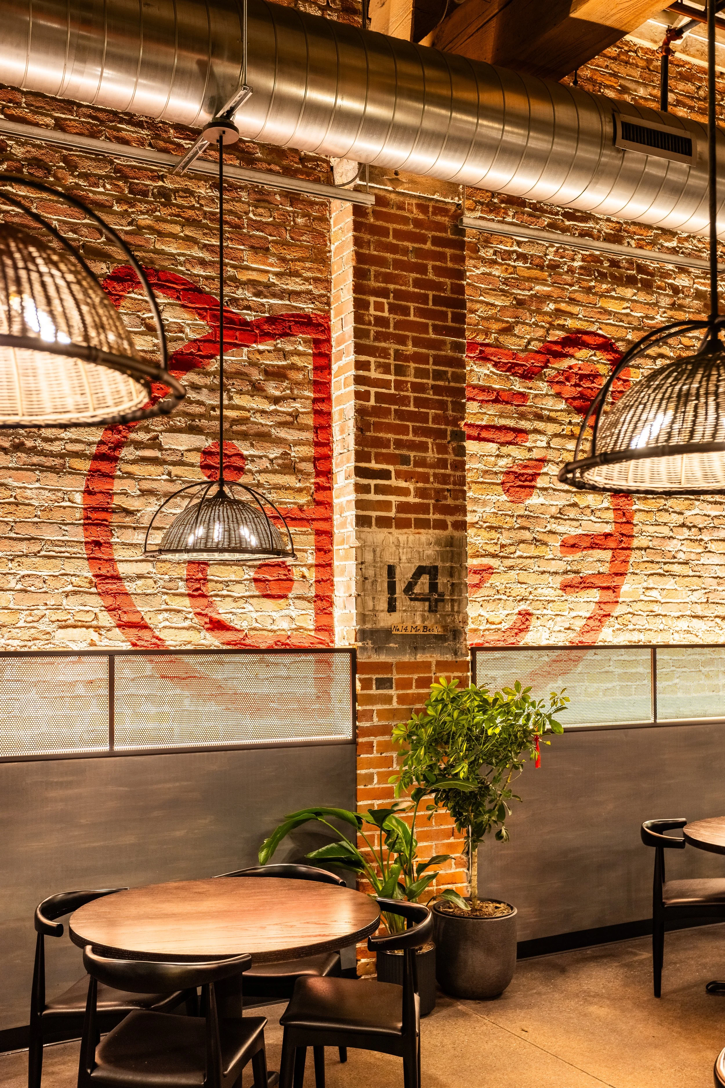 A brick wall with red spray-painted graffiti and the number 14, hanging industrial-style pendant lights, a small round wooden table with four black chairs, and a potted green plant in a cafe or restaurant interior.