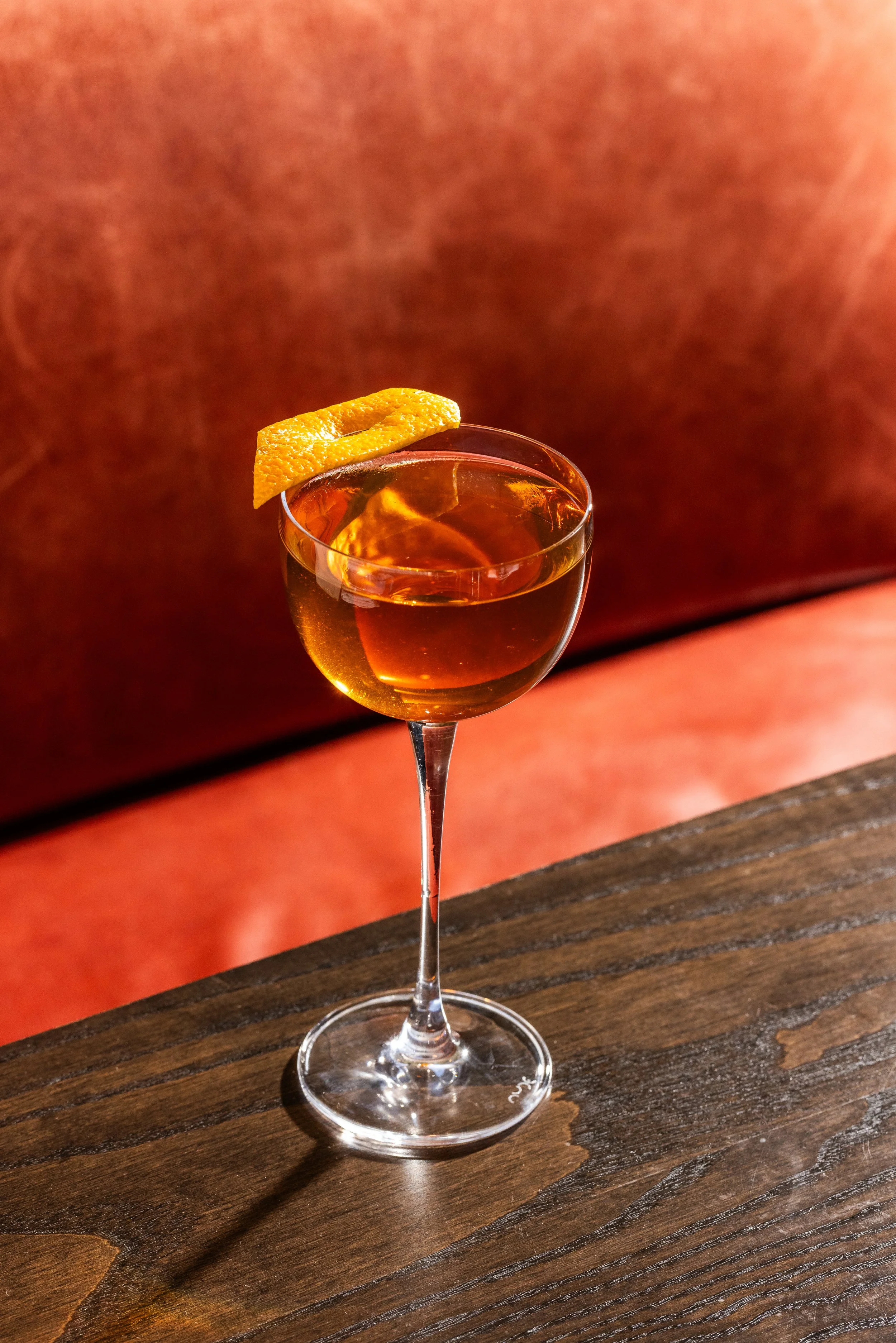 A cocktail glass filled with amber-colored drink, garnished with a twisted orange peel, placed on a wooden surface.