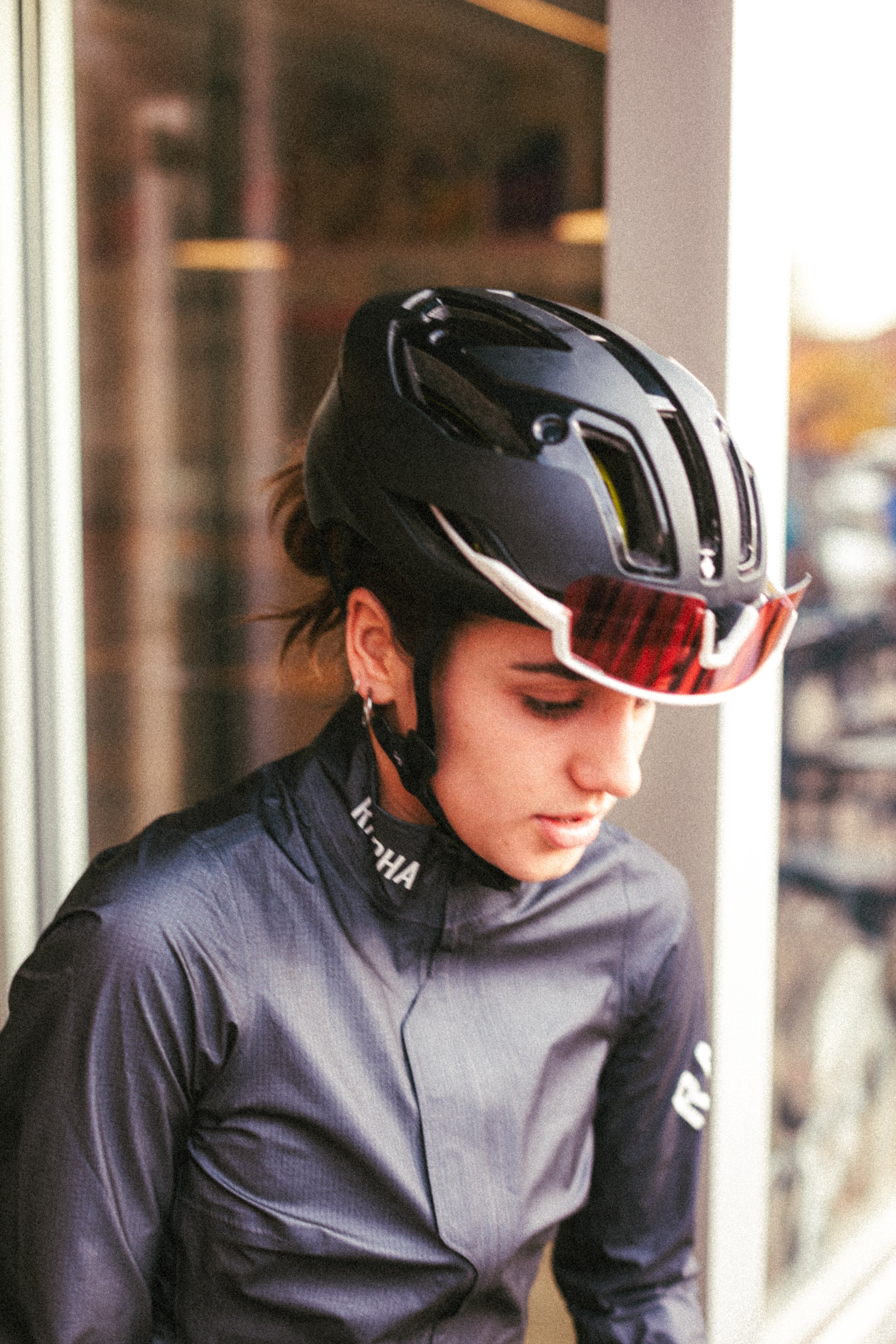 A young woman wearing a black bicycle helmet and sunglasses, dressed in a dark athletic jacket, standing indoors near a window.
