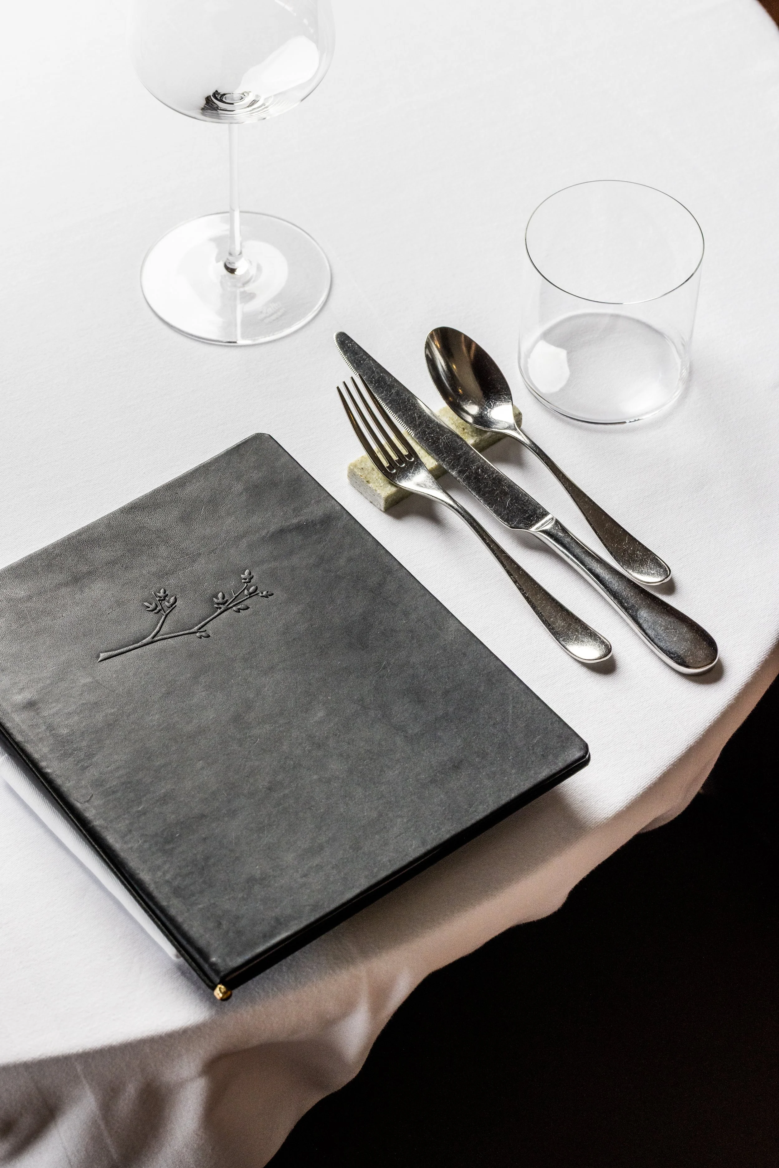 A table setting with a black menu featuring a branch illustration, silverware including a fork, knife, and spoon, a glass of water, an empty wine glass, and a clear glass on a white tablecloth.