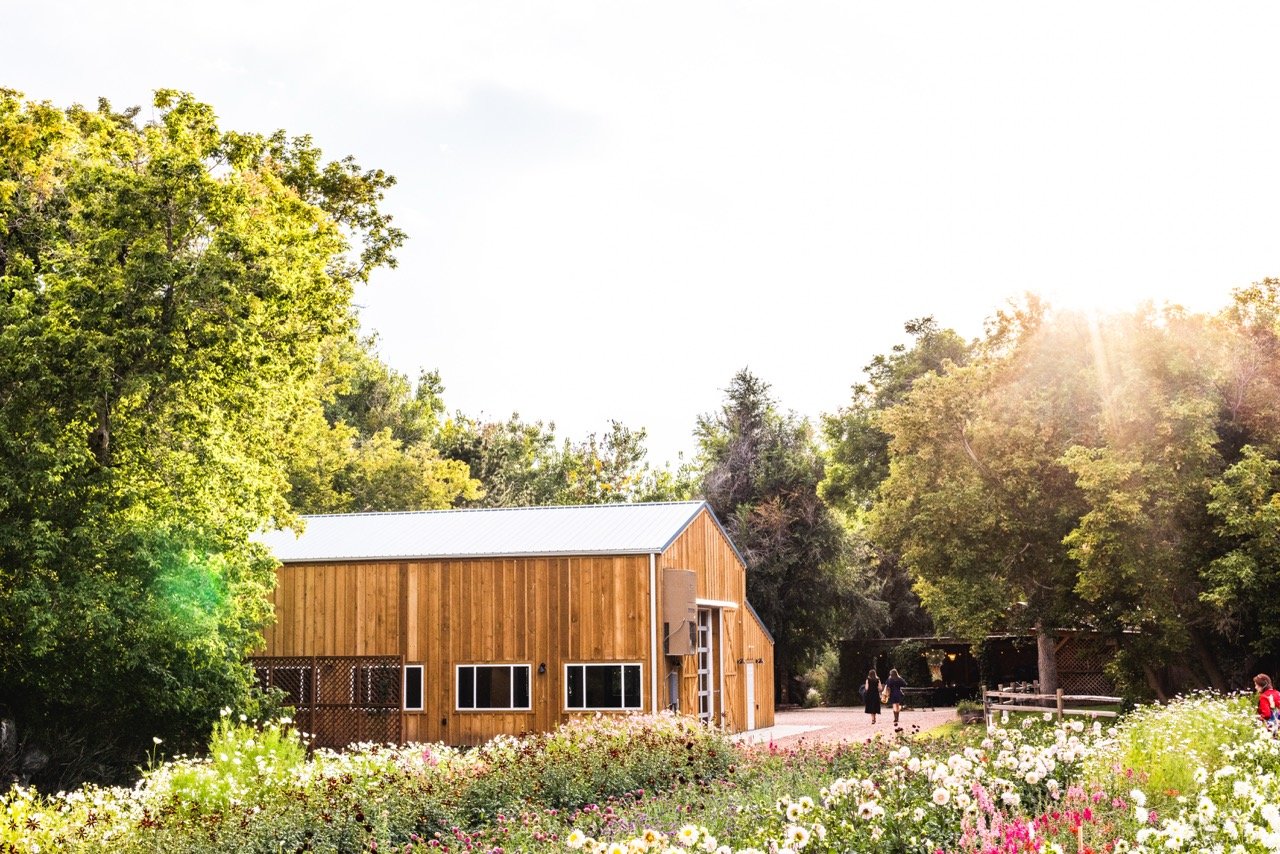 A wooden barn surrounded by colorful flowers and green trees, with a group of people walking nearby on a sunny day.