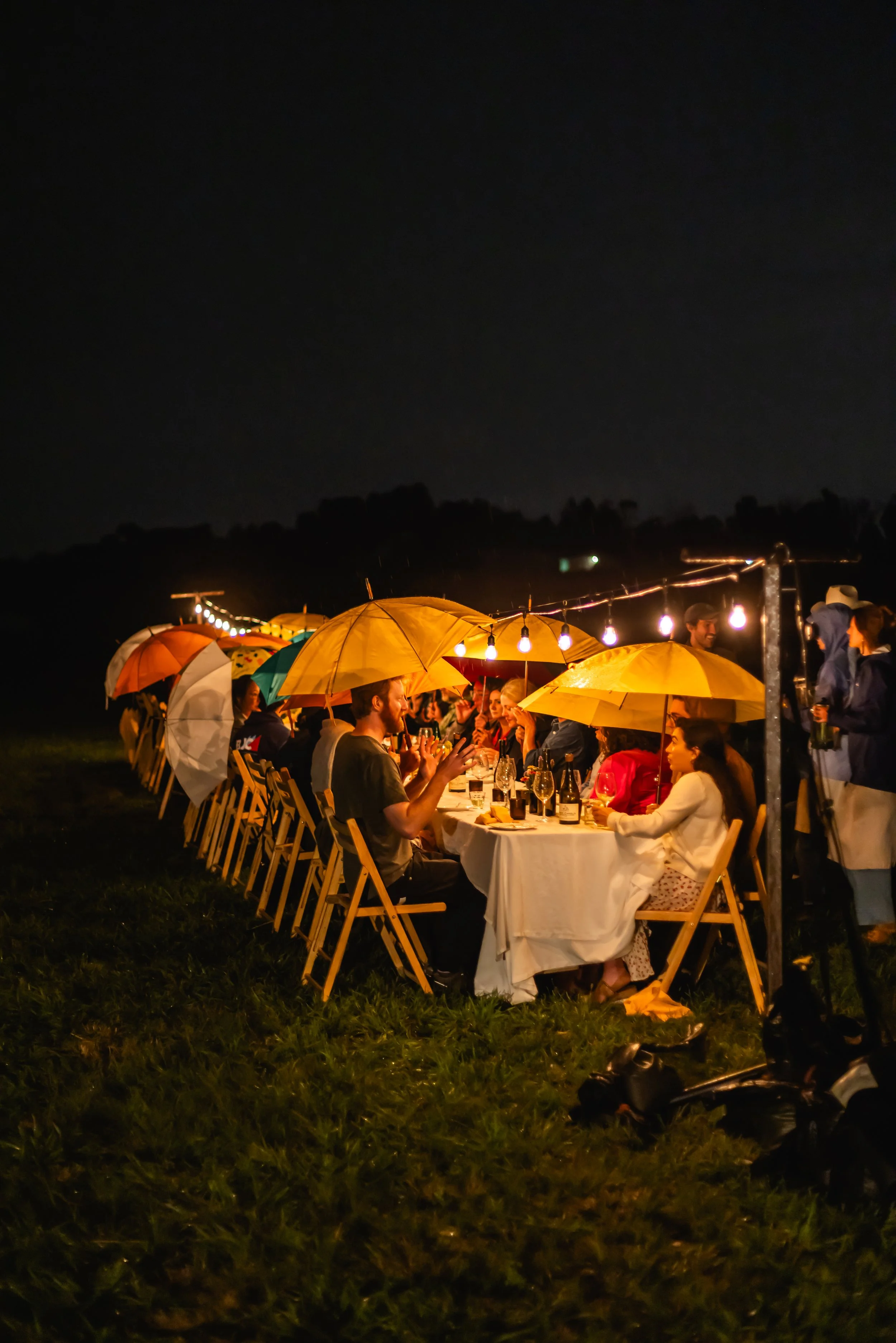 People dining outdoors at night under yellow umbrellas with string lights overhead.