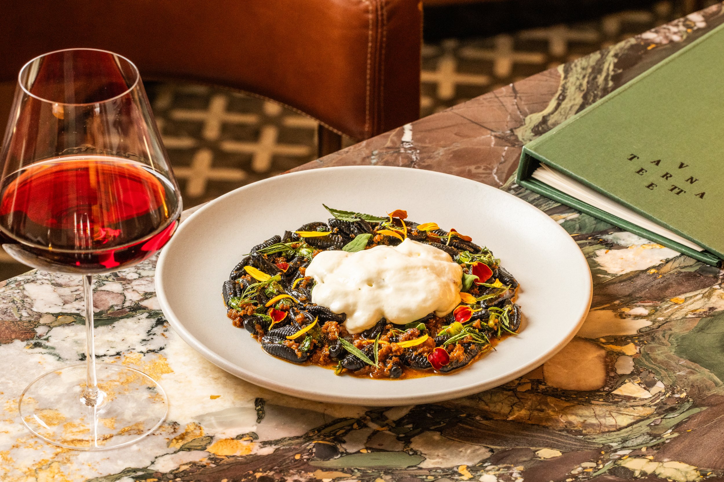 A plate of black truffle pasta topped with a dollop of cream, garnished with edible flowers and herbs, placed on a marble table with a glass of red wine and a green menu.