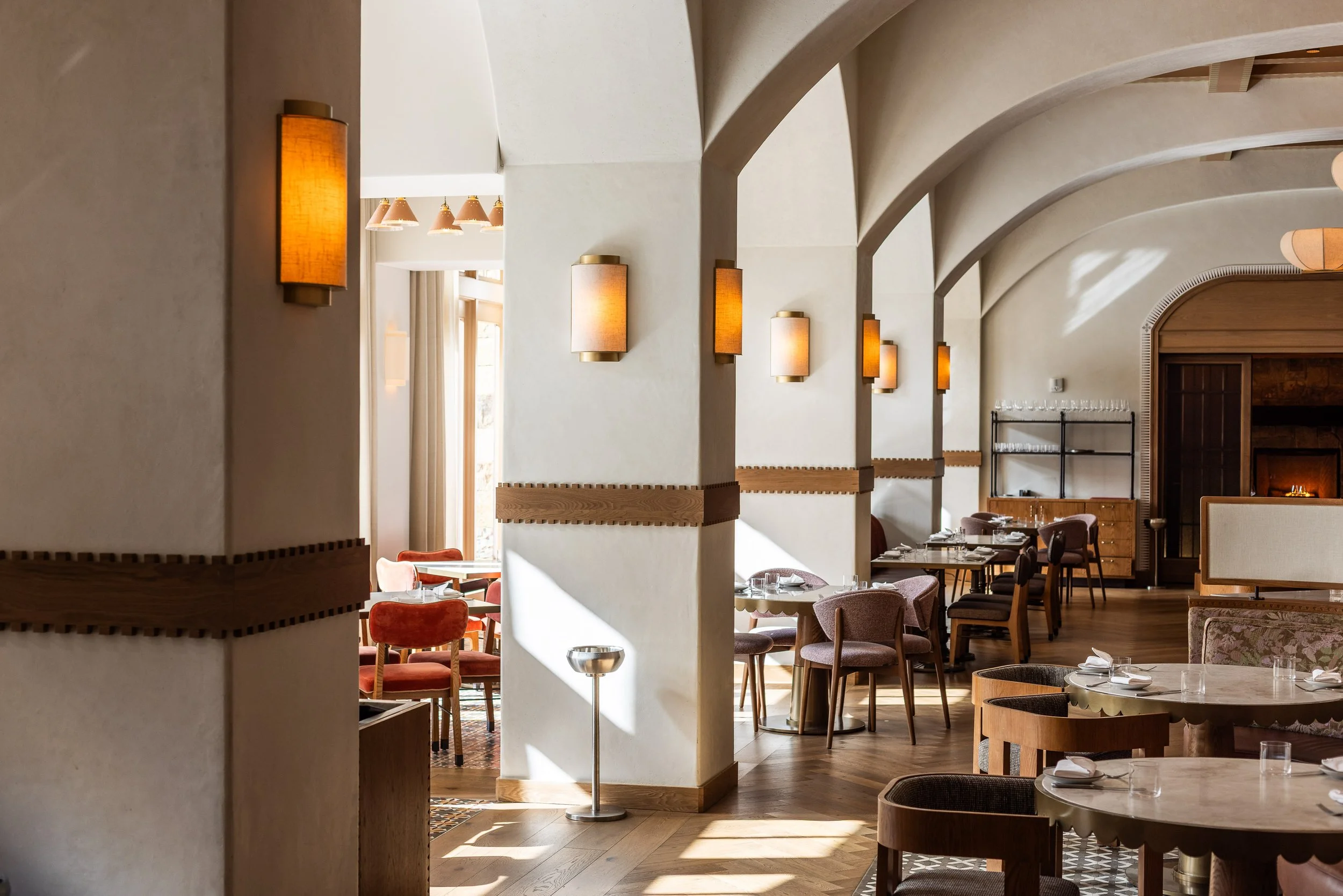 Interior of a restaurant with white walls, wooden accents, and warm lighting. Rows of tables and chairs are arranged with some tables set with plates, glasses, and napkins. Sunlight filters through windows creating bright patches on the wooden floor.