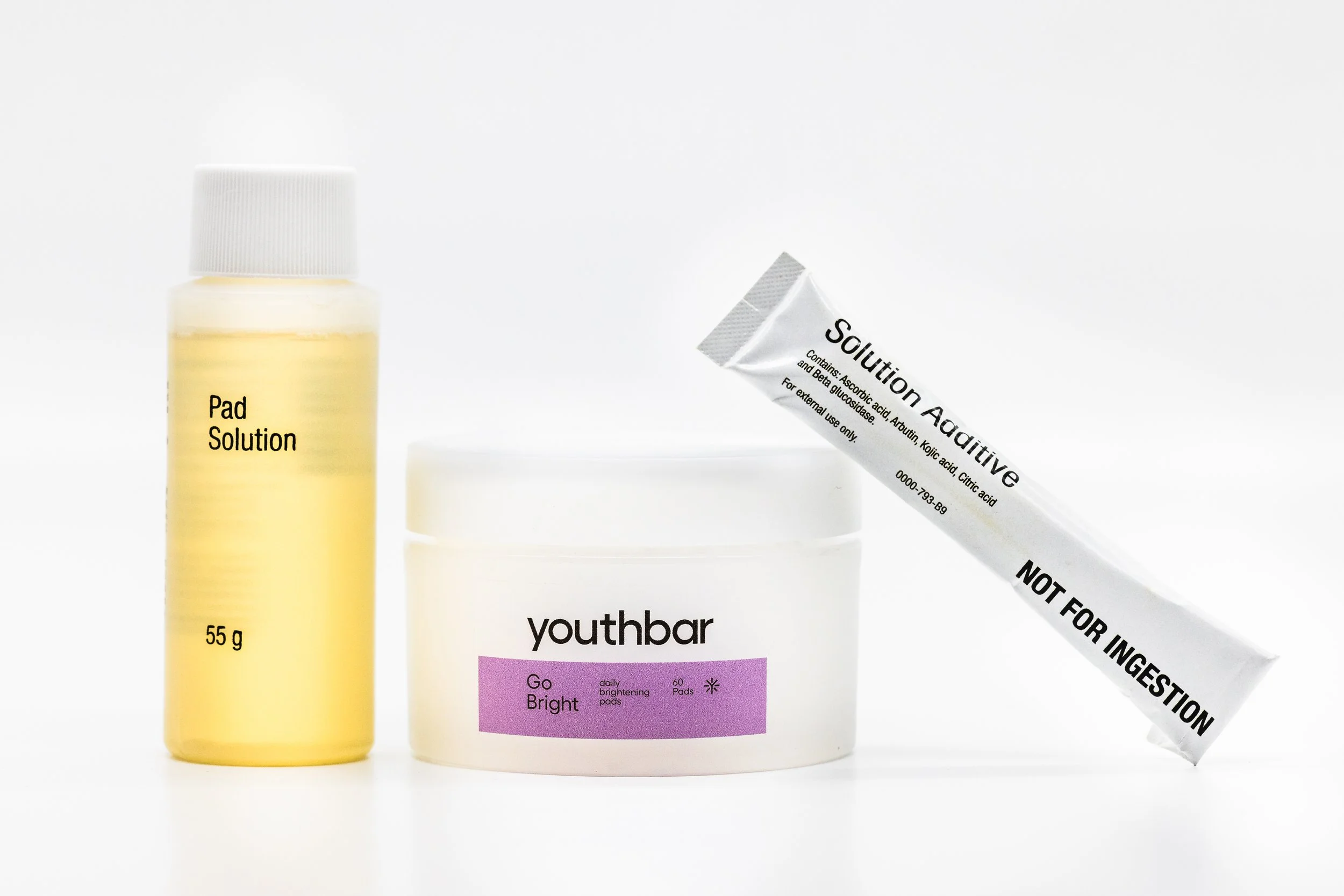 Collection of skincare products including a yellow bottle labeled 'Pad Solution 55g', a white container labeled 'youthbar Go Bright daily brightening pads', and a white tube labeled 'Solution Additive NOT FOR INGESTION' with black text.