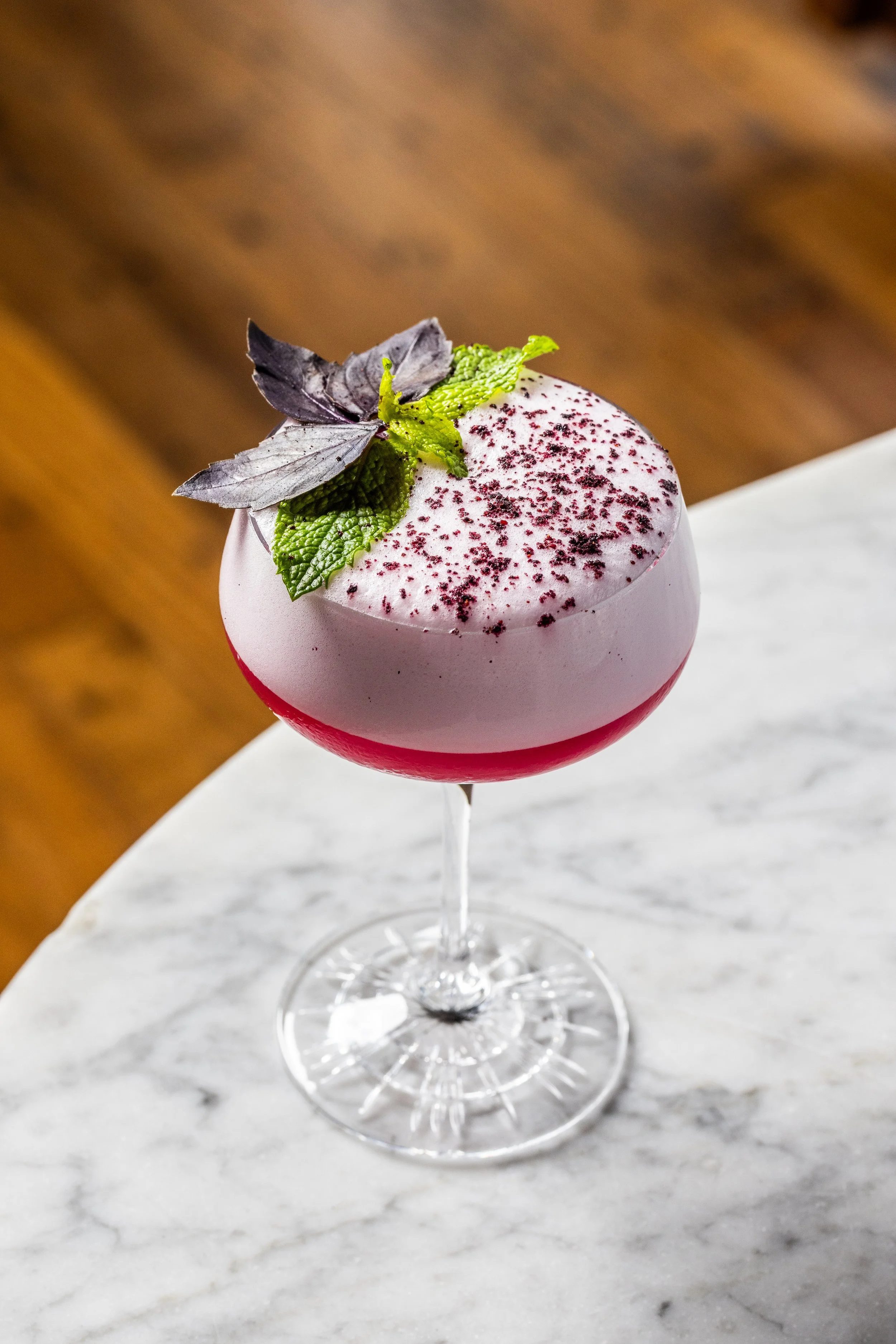 A cocktail in a coupe glass with a pink and white layered topping, garnished with mint and purple basil leaves, and sprinkled with red powder.