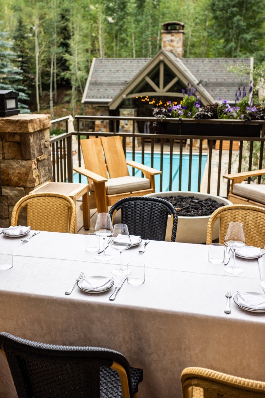 Outdoor dining table set with plates, utensils, and glasses, overlooking a deck with chairs, a fire pit, and a swimming pool, surrounded by trees and a house with a stone chimney.