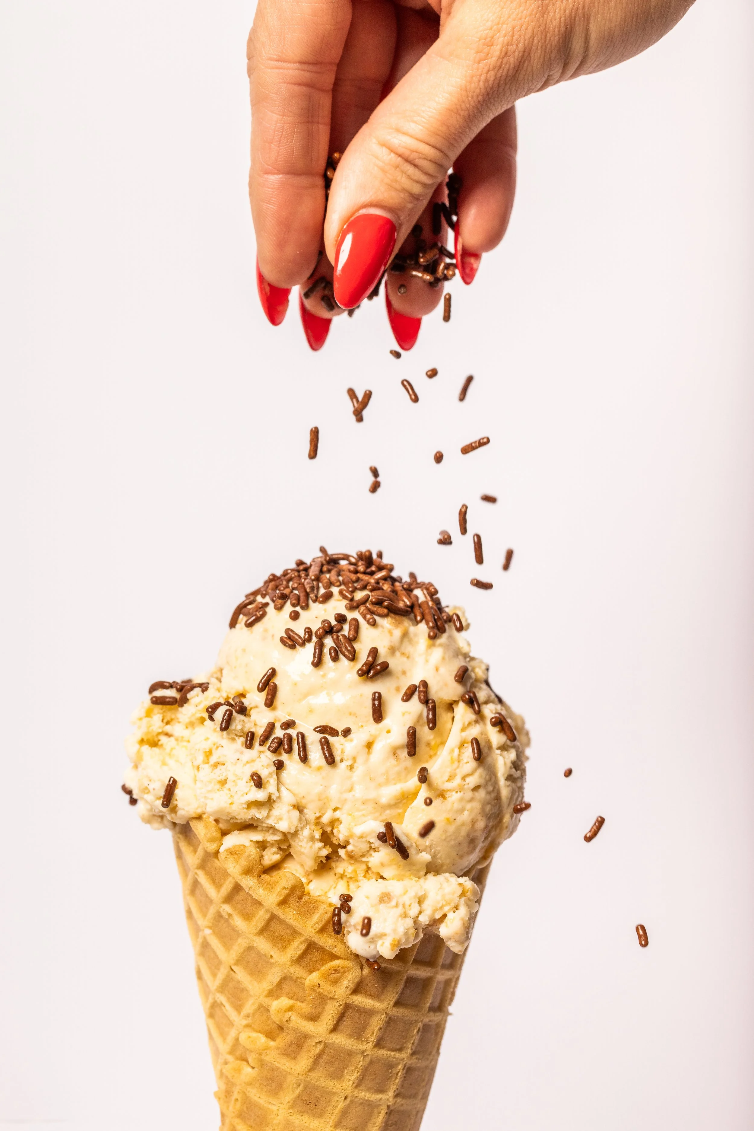 Hand with red painted nails sprinkling chocolate sprinkles onto a scoop of vanilla ice cream in a waffle cone.