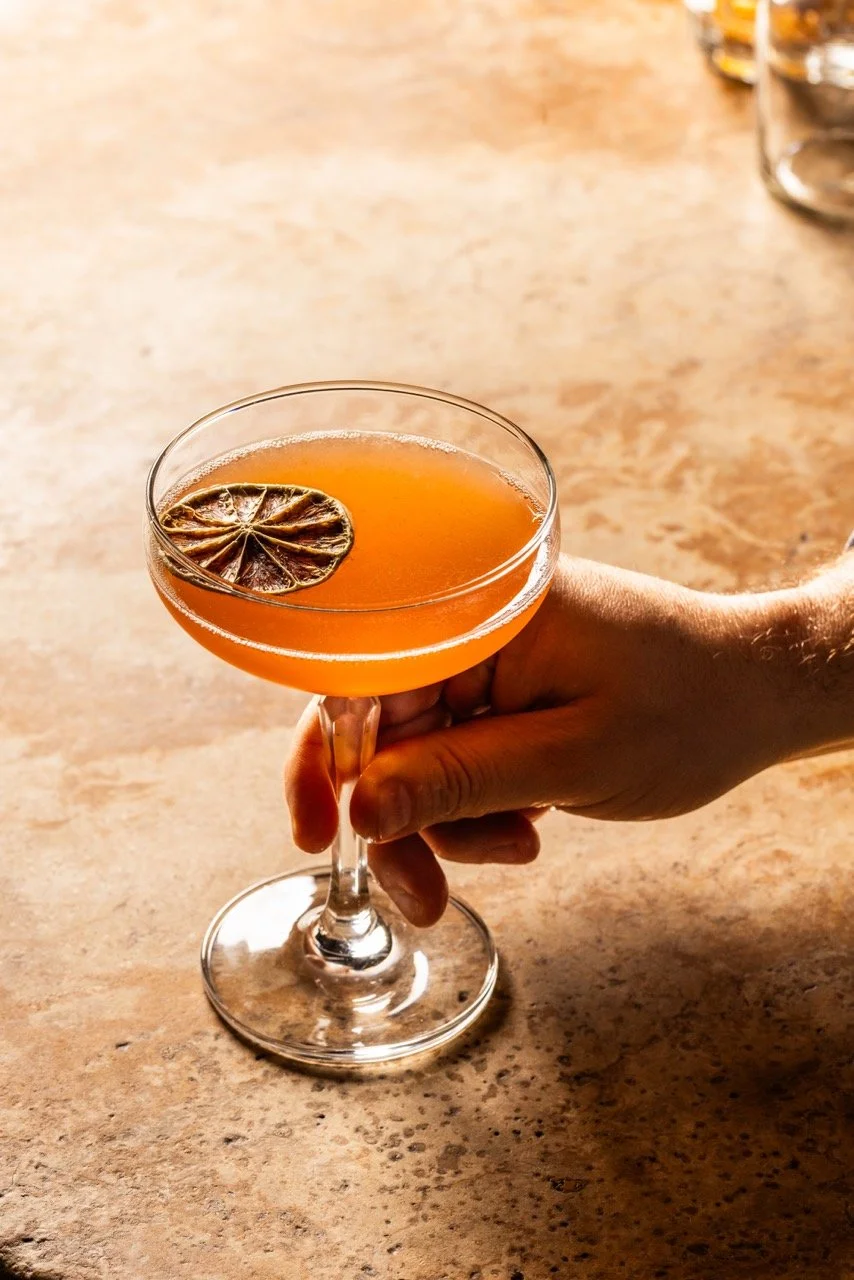 A hand holding a cocktail glass with an orange-colored drink and a dried lemon slice garnish, on a wooden surface.
