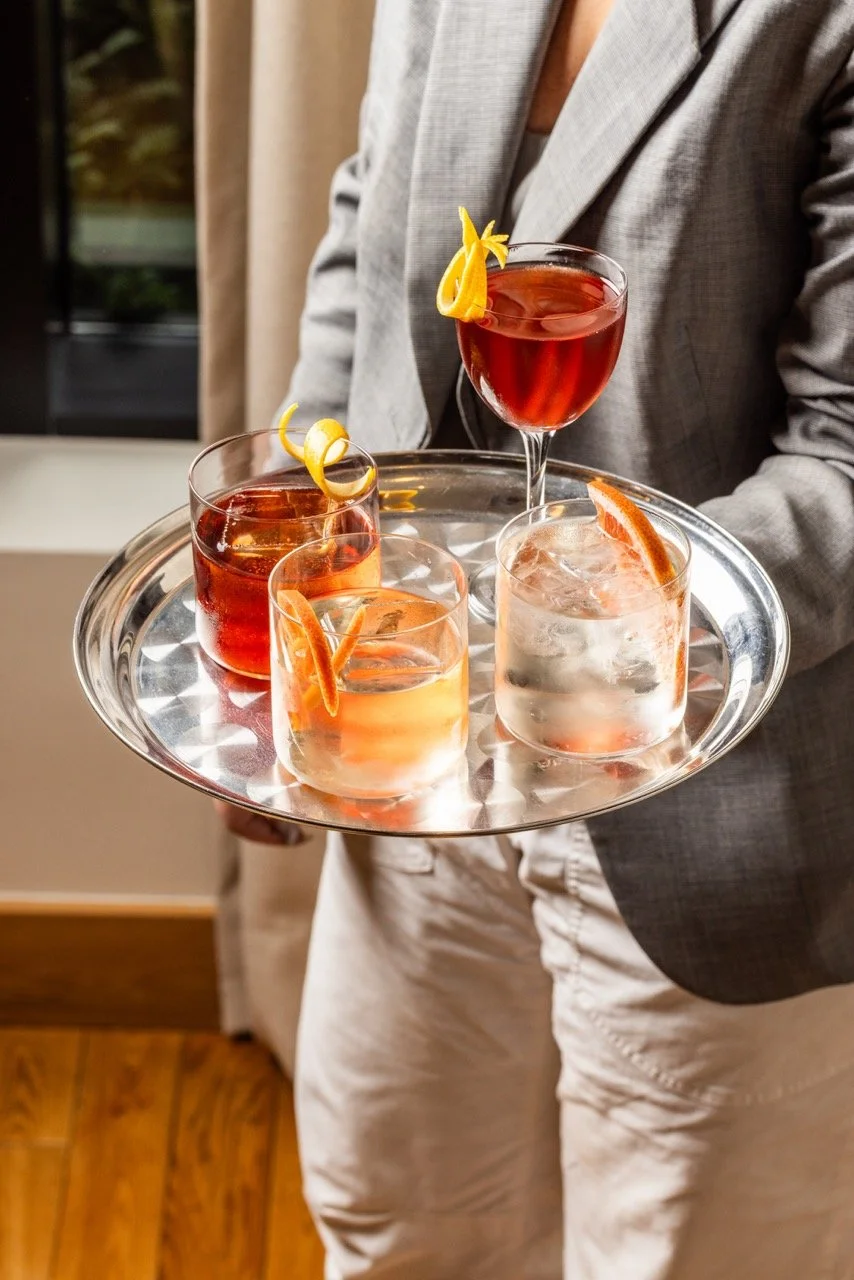 A waiter holding a round silver tray with four cocktails garnished with citrus peels and slices, in a room with wooden flooring and a window.