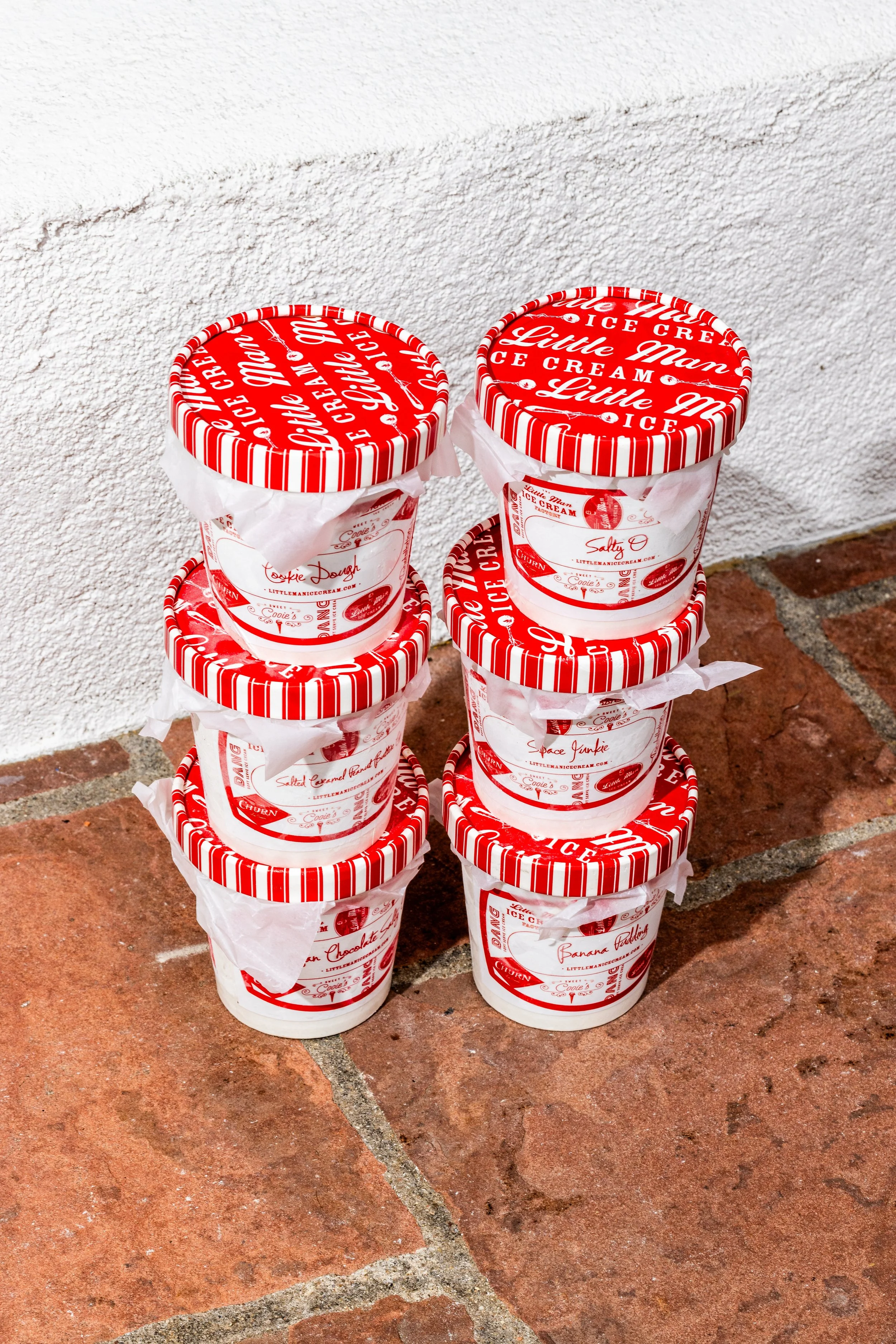 Stacks of six Little Man Ice Cream containers with red and white striped lids, arranged on a brick sidewalk next to a white wall.