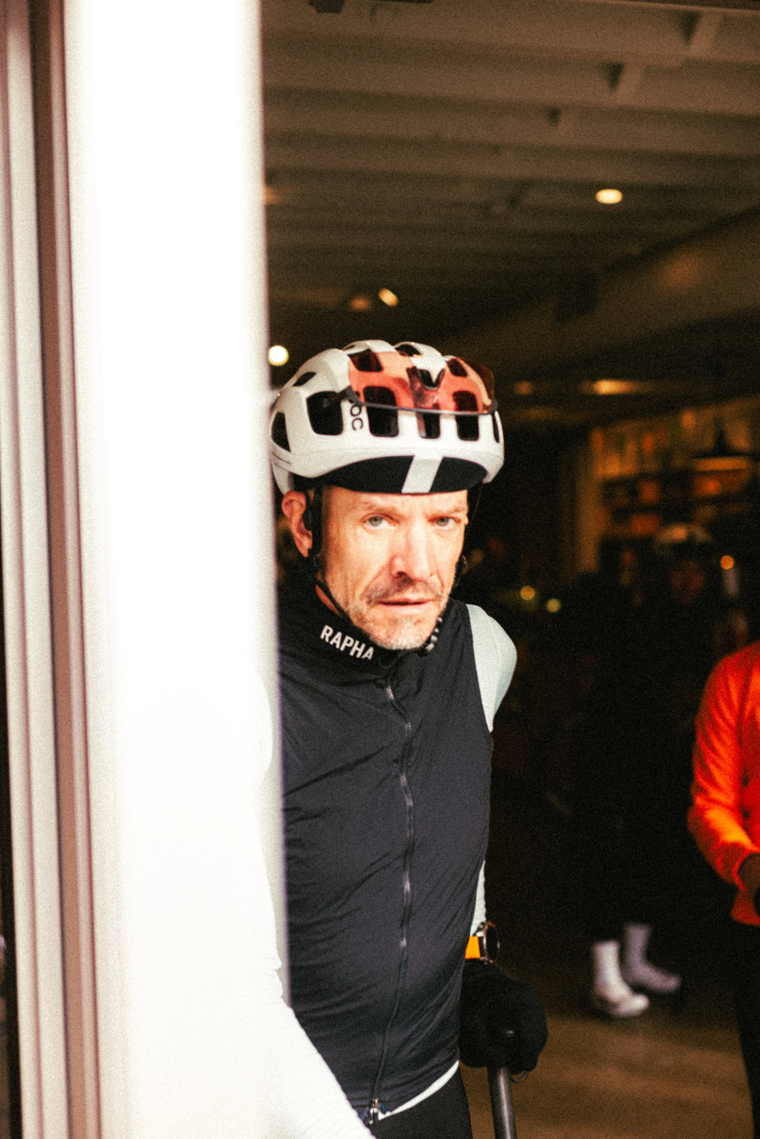 A middle-aged man wearing a bicycle helmet peering around a corner, dressed in cycling gear, indoors.