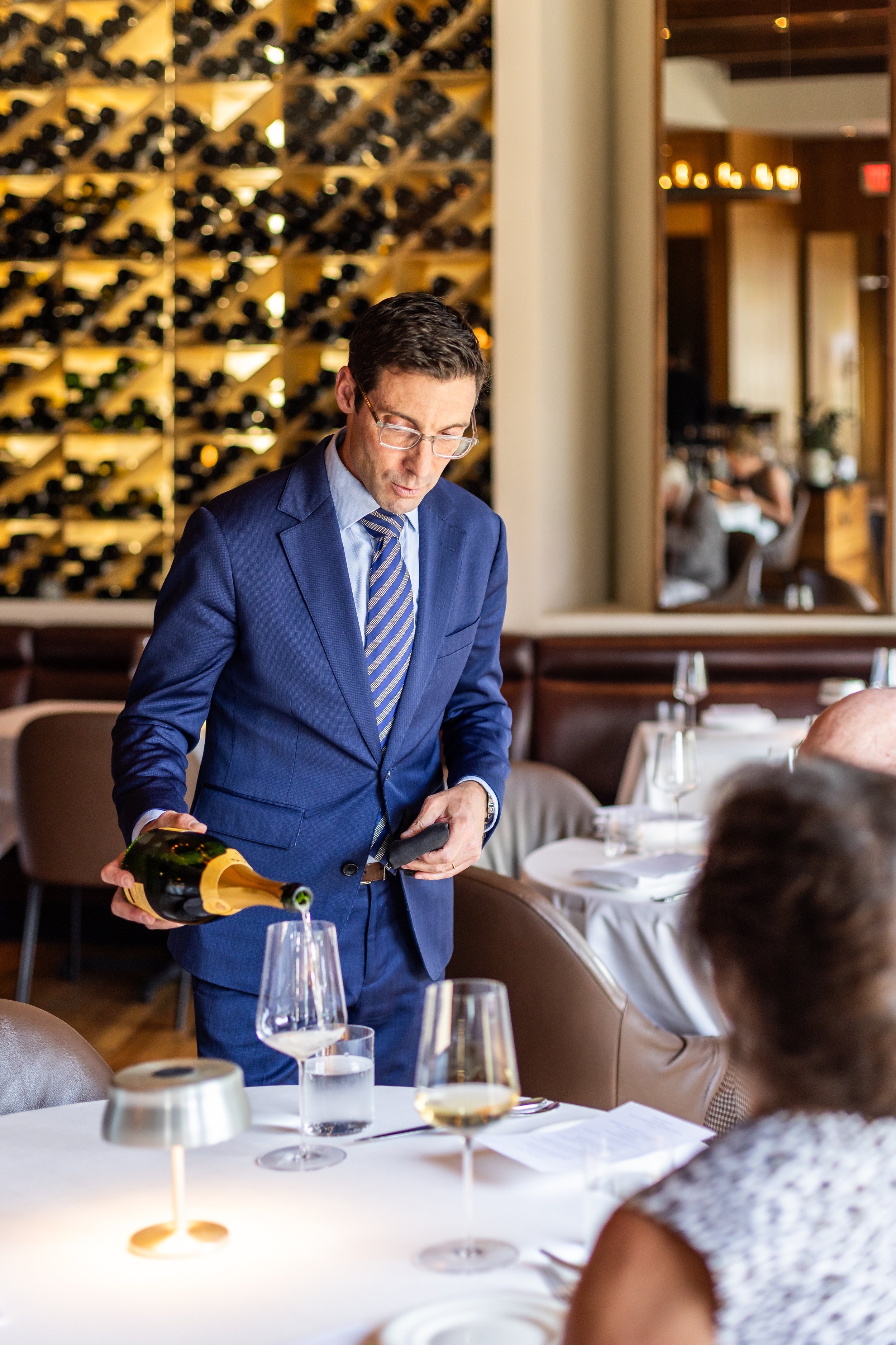 A man in a blue suit is pouring white wine into a glass at a restaurant table, with other tables and wine racks in the background.