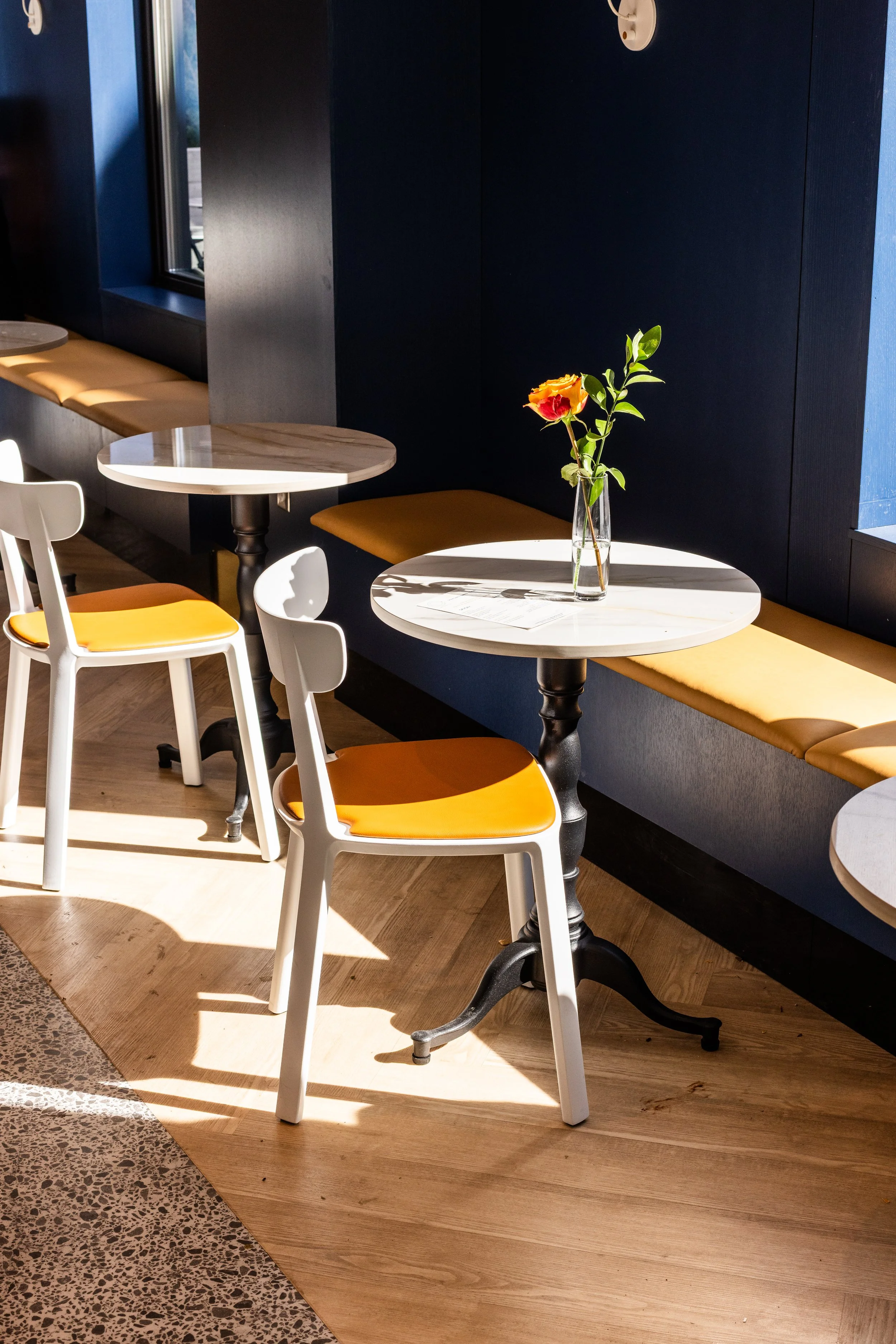 Sunlit cafe with white marble-top tables, white and yellow chairs, and a Venosa-filled vase on one table.