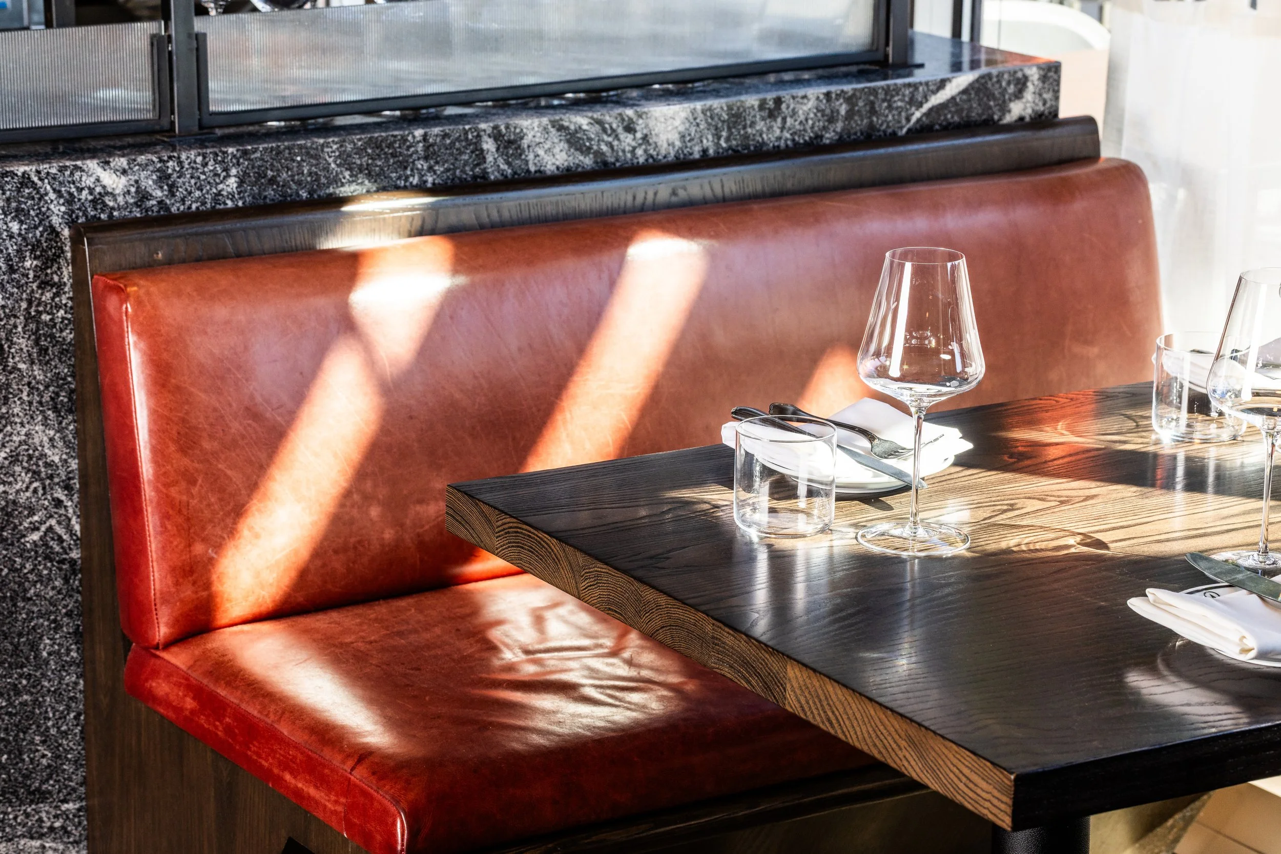 A restaurant booth with a red leather seat and a wooden table set with wine glasses, water glasses, plates, and cutlery, with sunlight casting shadows on the booth.