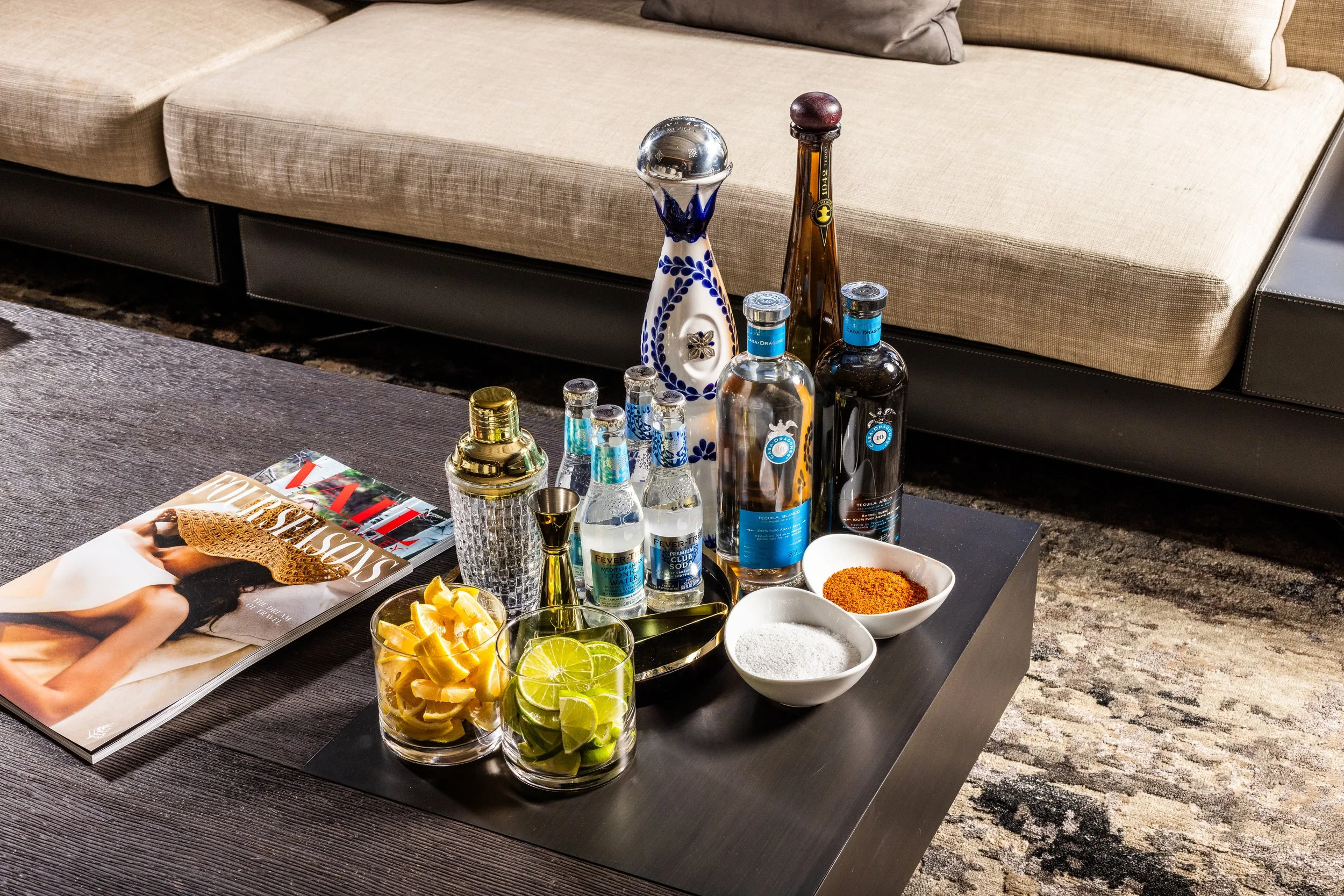 A coffee table with various bottles of alcohol, glasses, bowls of salt and chili powder, and two glasses with lemon and lime slices, in front of a beige sofa with cushions, in a living room.