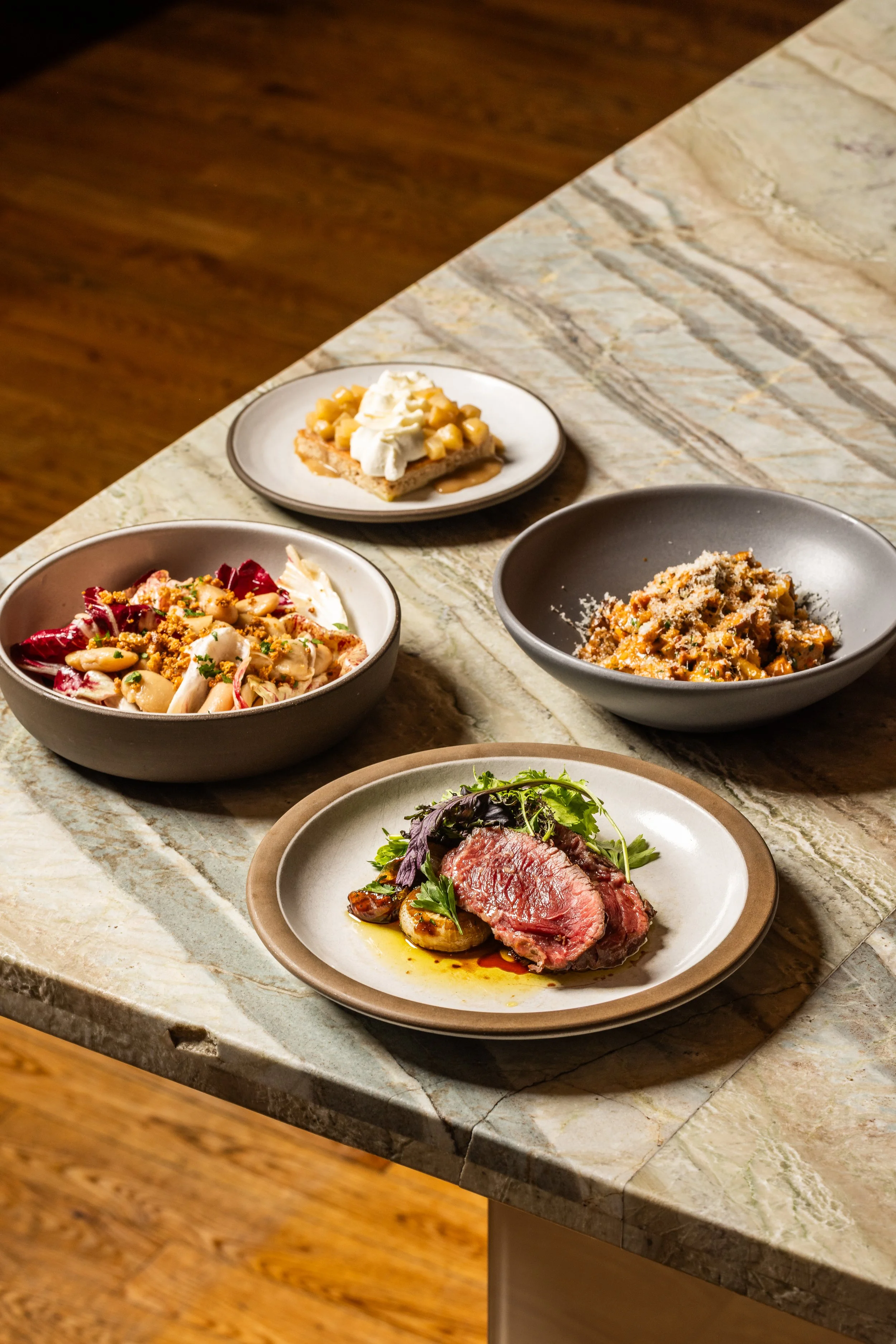 A marble kitchen countertop with five dishes, including a plate of sliced cooked beef with greens, a bowl of vegetable salad, a bowl of pasta with sauce, a dessert with whipped cream and corn, and another dessert with biscuit base and whipped topping