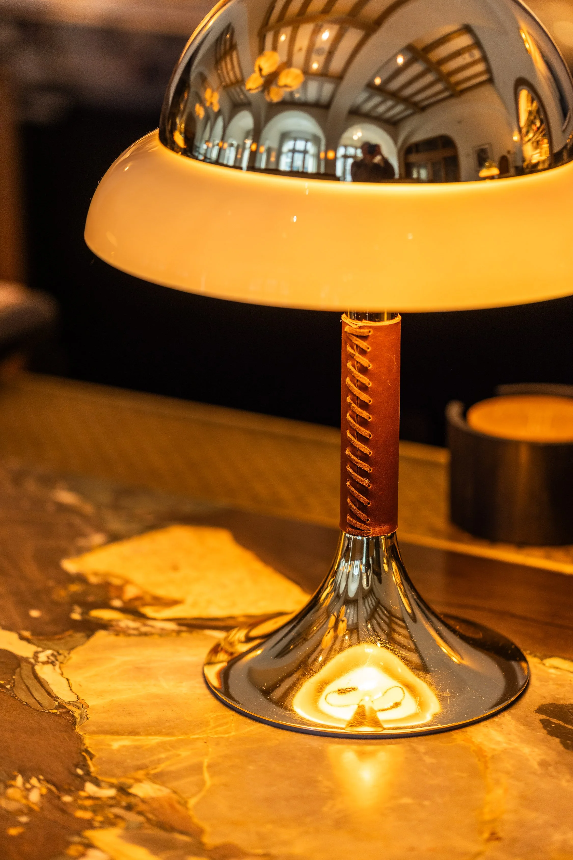 Close-up of a modern table lamp with a shiny metallic base and a beige lampshade, reflecting the interior of a room with high ceilings, large windows, and hanging lamps.