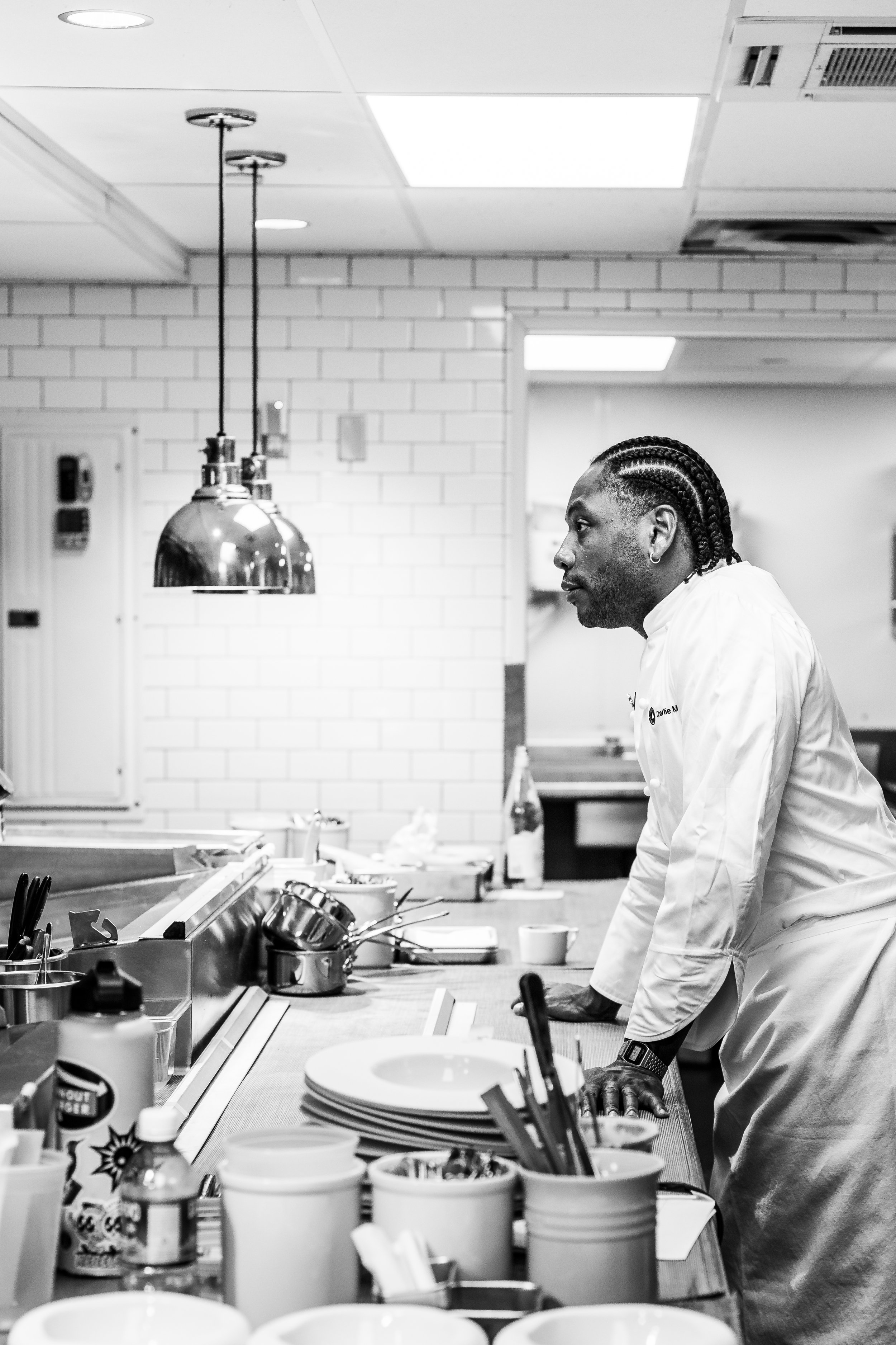 A man in a chef's coat leaning on a kitchen counter in a restaurant or commercial kitchen.