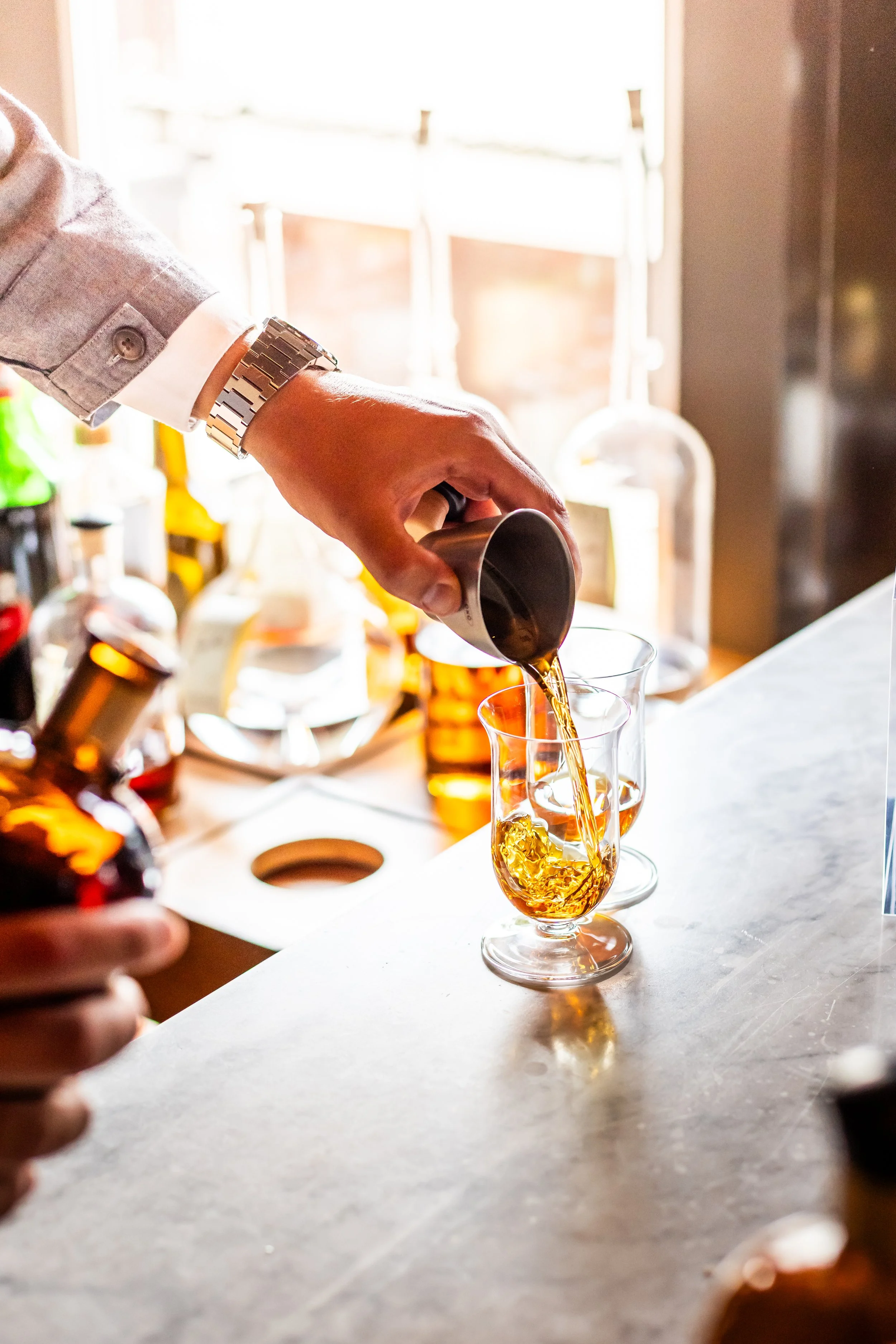 Person pouring a drink into a glass at a bar with various bottles in the background.