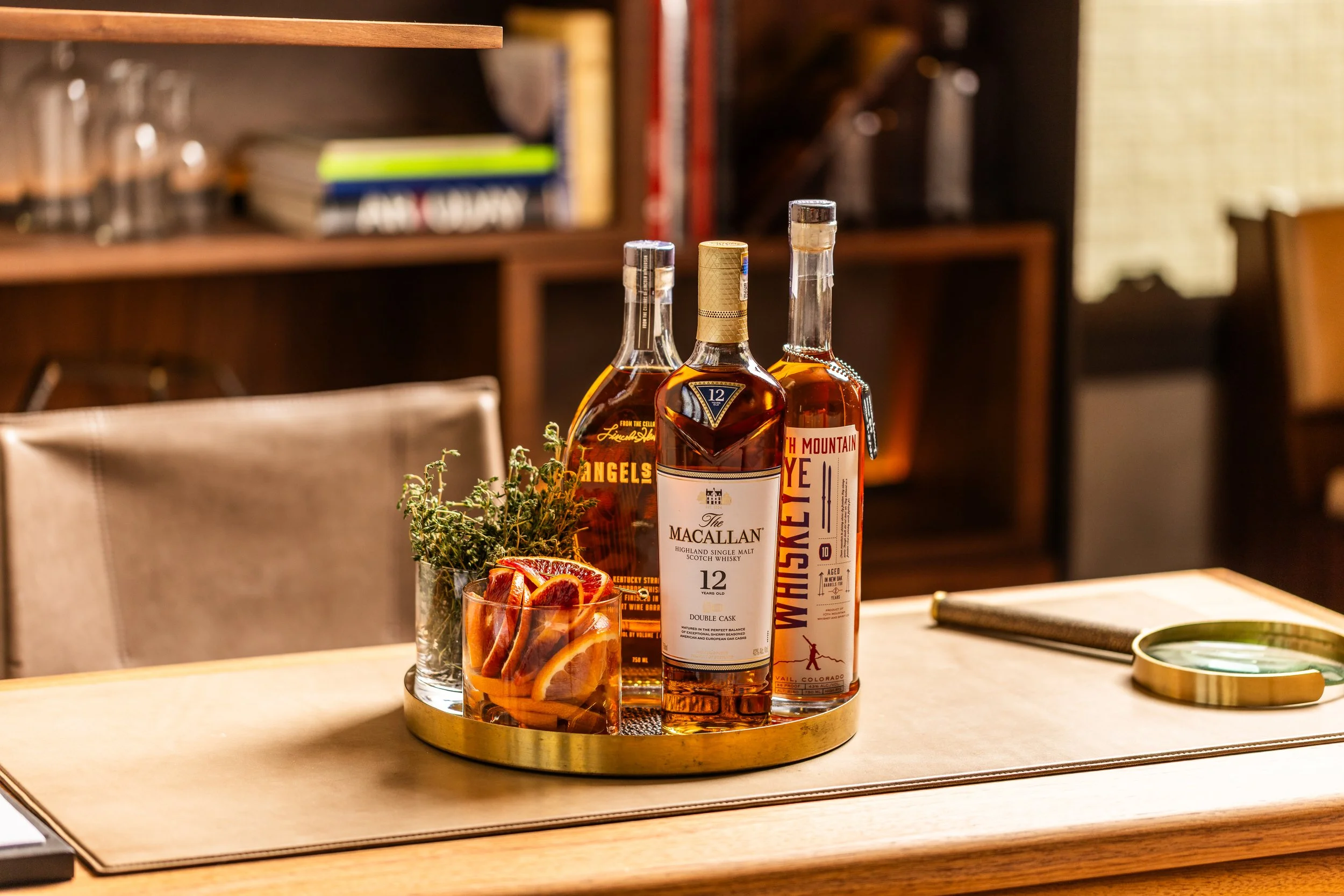 Three bottles of whiskey on a gold tray, along with a glass filled with slices of orange and herbs, and a small dish with lemon slices, on a wooden table in a cozy, well-lit room.