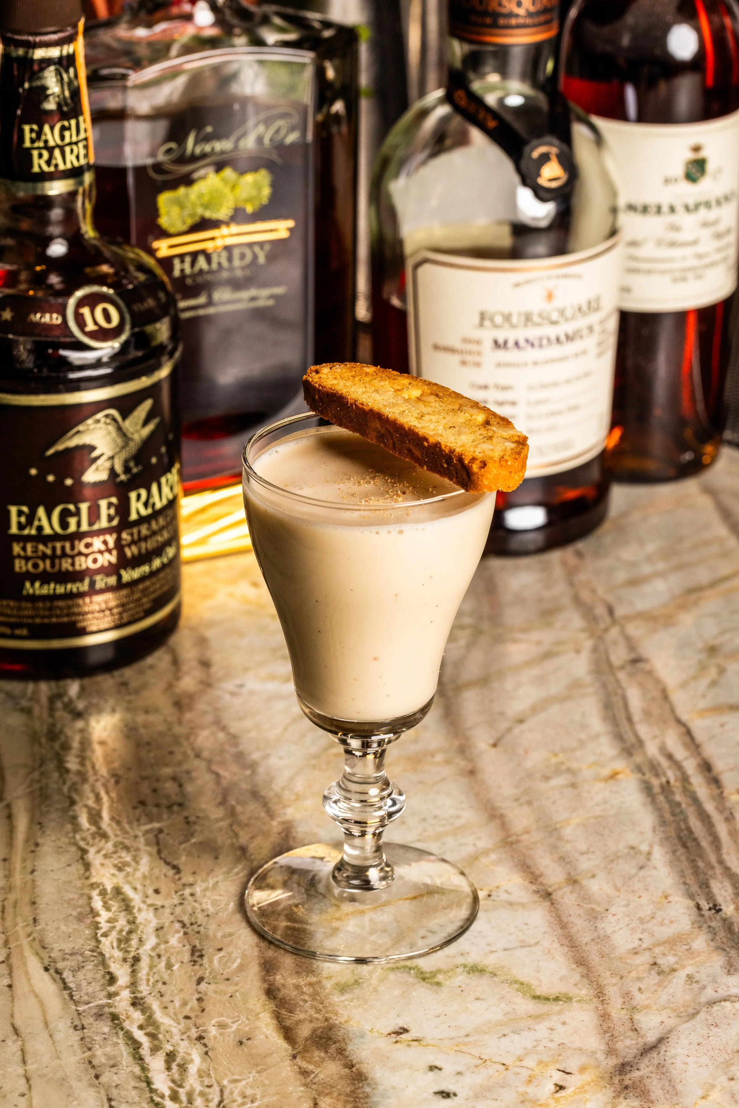 A creamy cocktail garnished with a toasted bread slice on a marble countertop, with bottles of whiskey in the background.