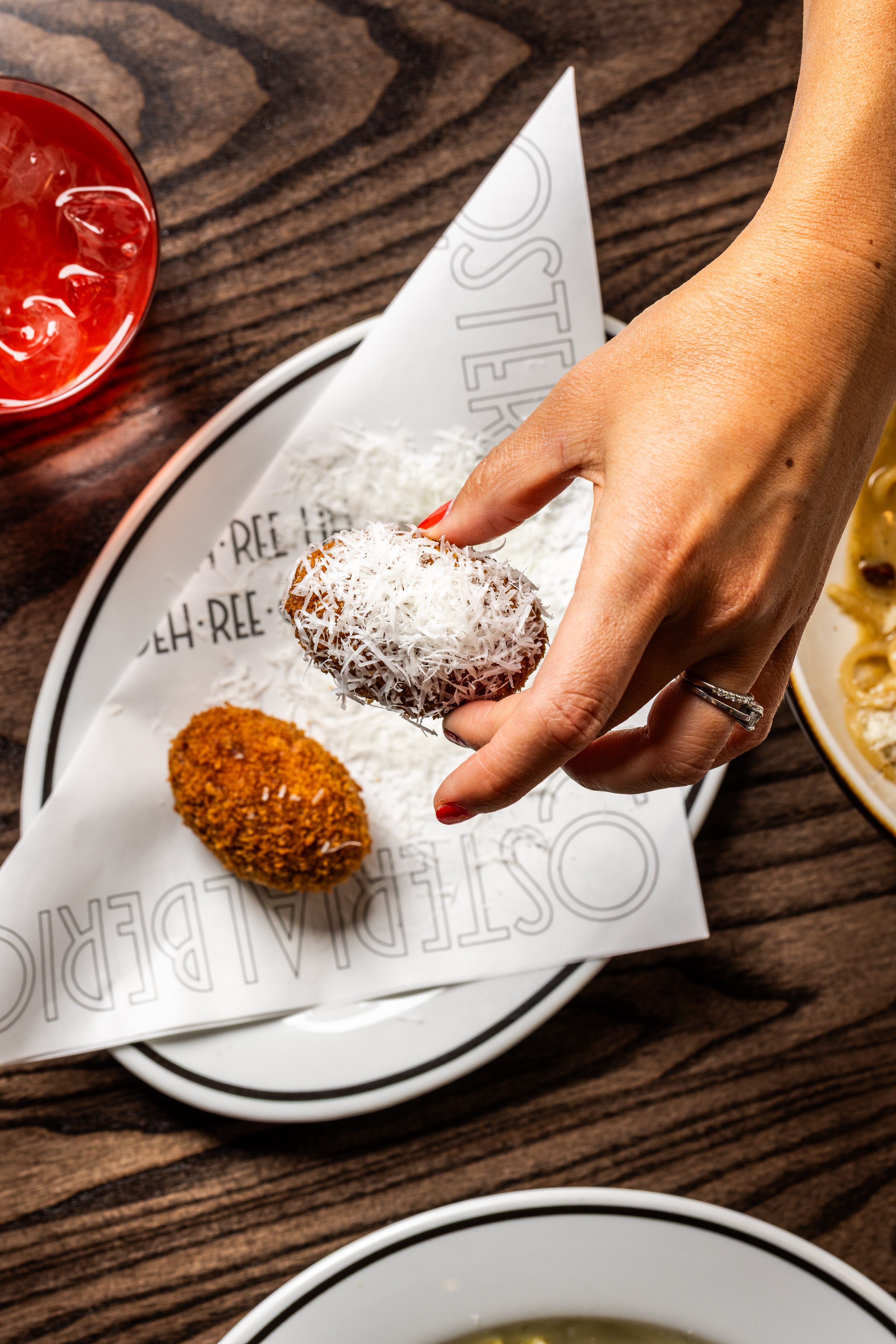 Person holding a coconut-covered dessert with shredded coconut, with another similar dessert on a white plate, on a wooden table.