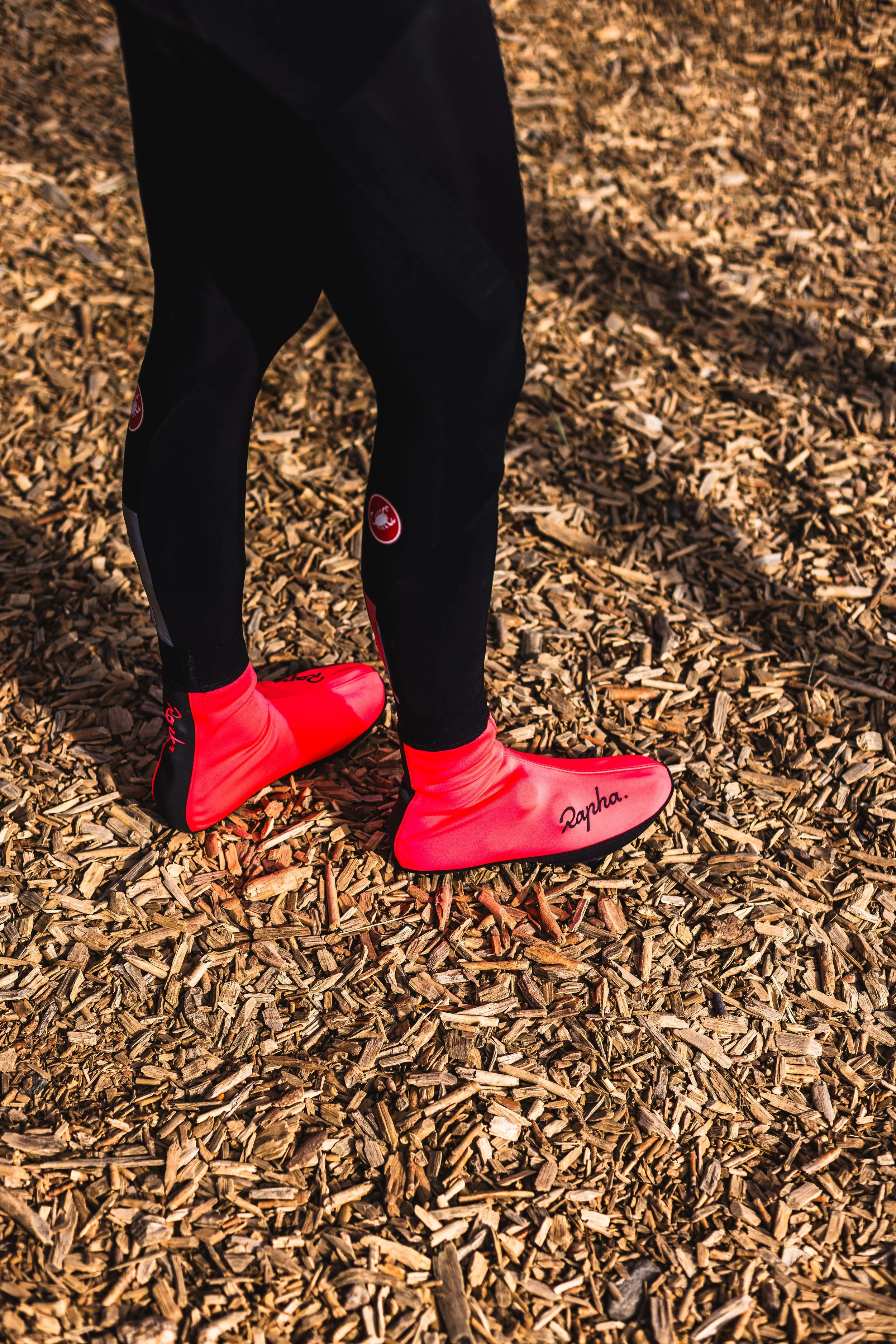 A person wearing black athletic leggings with a Rapha logo and bright red overshoes standing on wood chips.