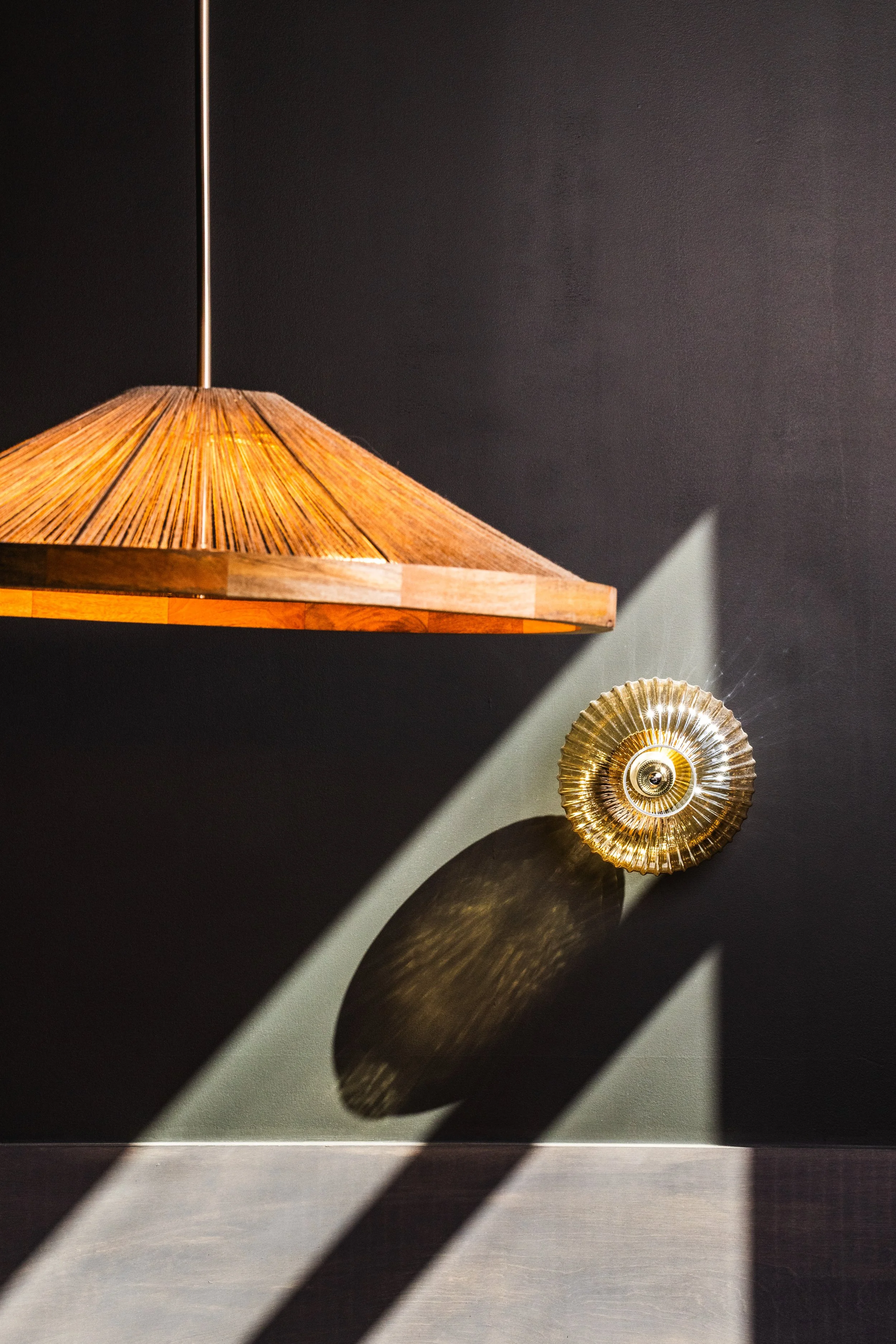 A wooden hanging lamp with a conical shade and a shiny, round, gold decorative piece on a black wall, casting shadows.