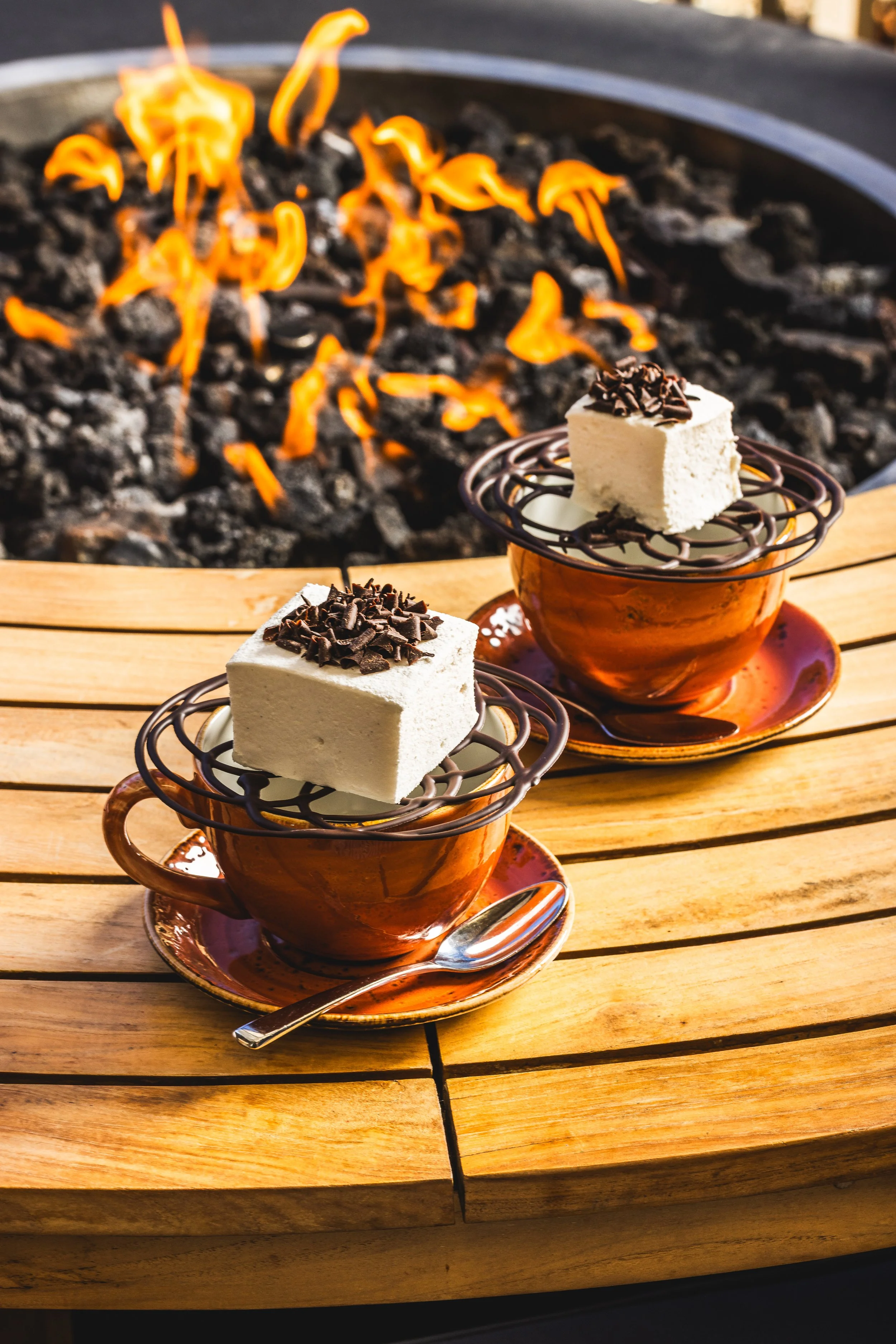 Two cups of coffee with slices of cheesecake topped with chocolate shavings, placed on a wooden table with a campfire in the background.