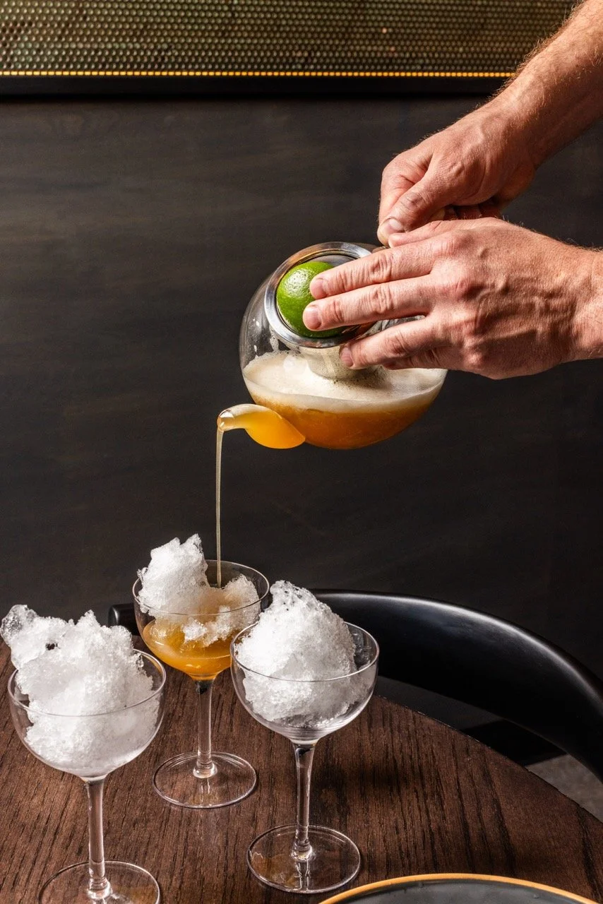 A person pouring a beverage from a jar over shaved ice in glasses, with a lime lodged in the jar's opening.