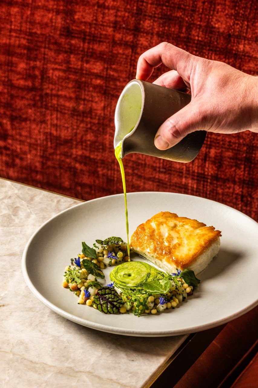 A hand pouring green sauce over a plated dish with a piece of seared fish, a swirl of sauce, and colorful salad garnishes on a white plate.