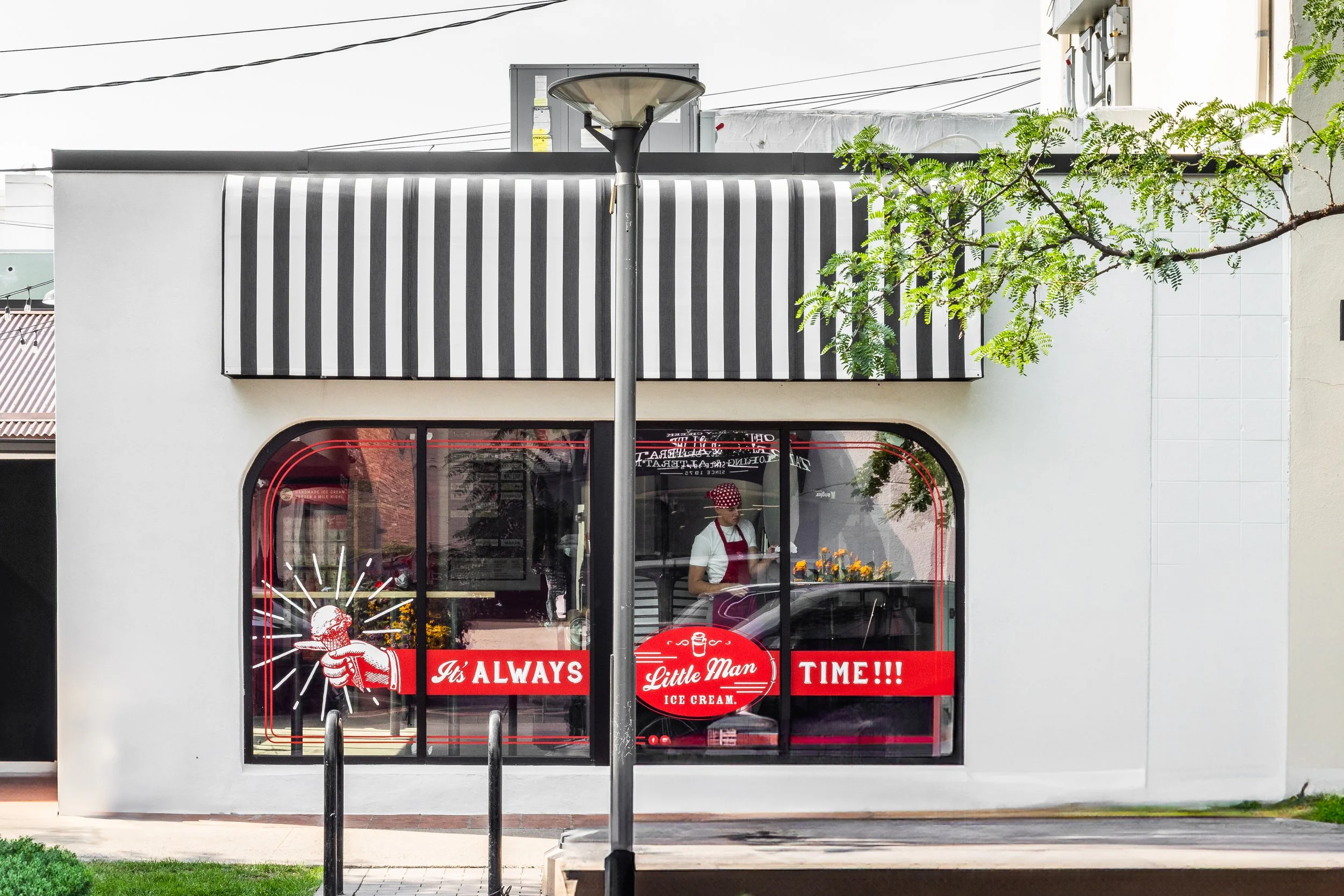 Front of a small ice cream shop with a large window, red and white signage, and a sign that reads 'It's ALWAYS Little Man ICE CREAM TIME!!!' A person in a red cap and apron is inside.