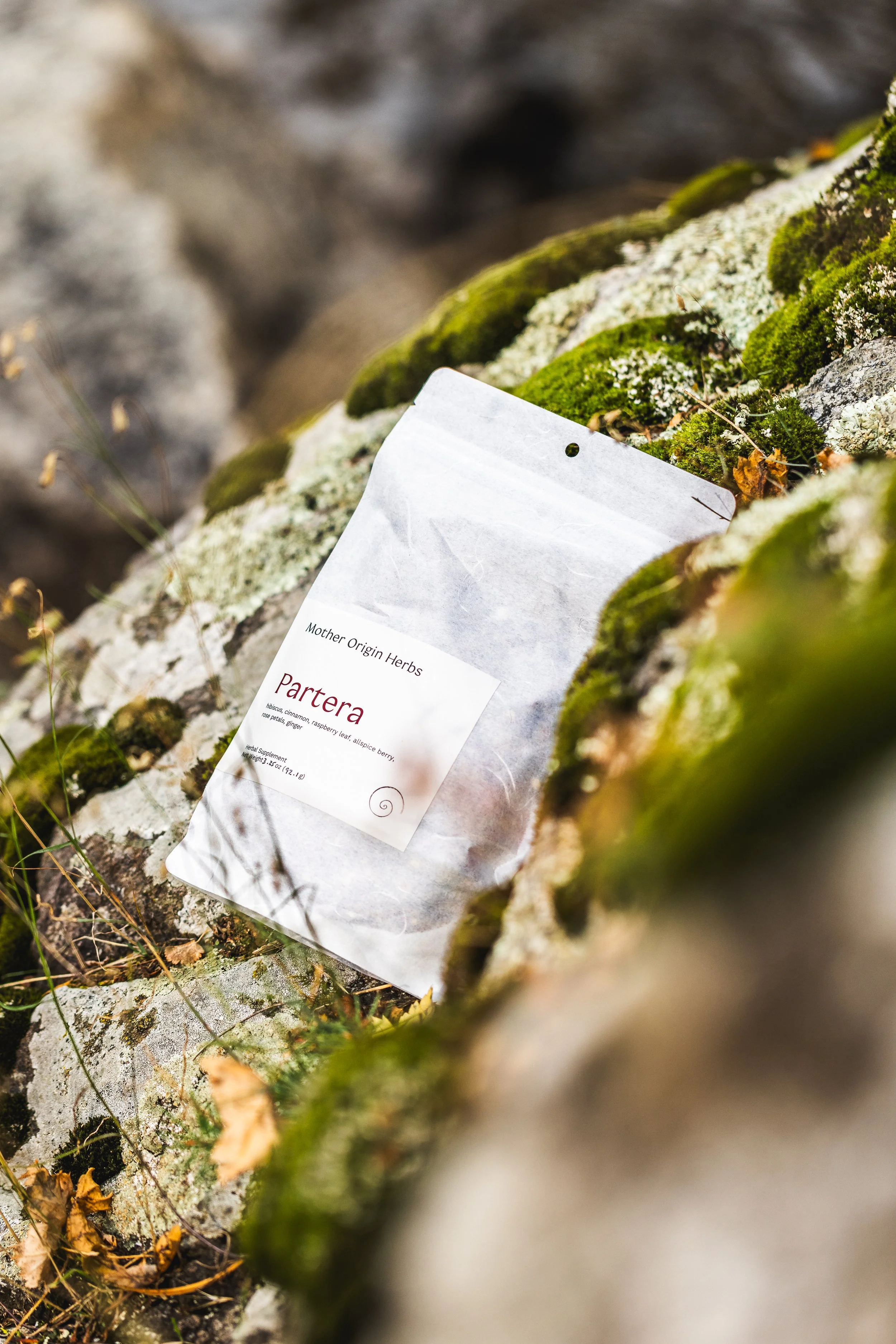 A packet of dried herbs labeled 'Mother Origin Herbs' on a moss-covered rock outdoors.