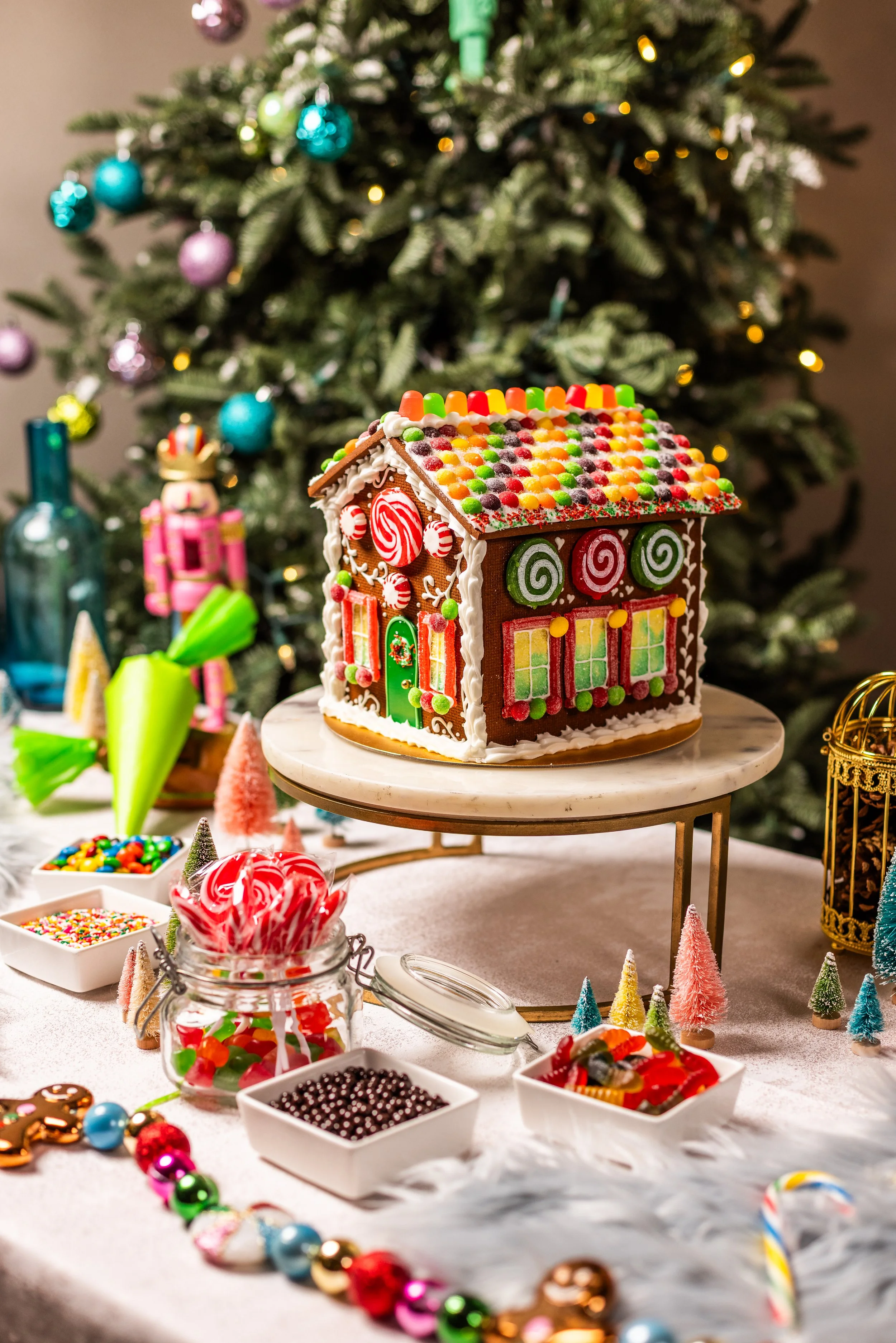Decorated gingerbread house with colorful candies in front of a decorated Christmas tree.