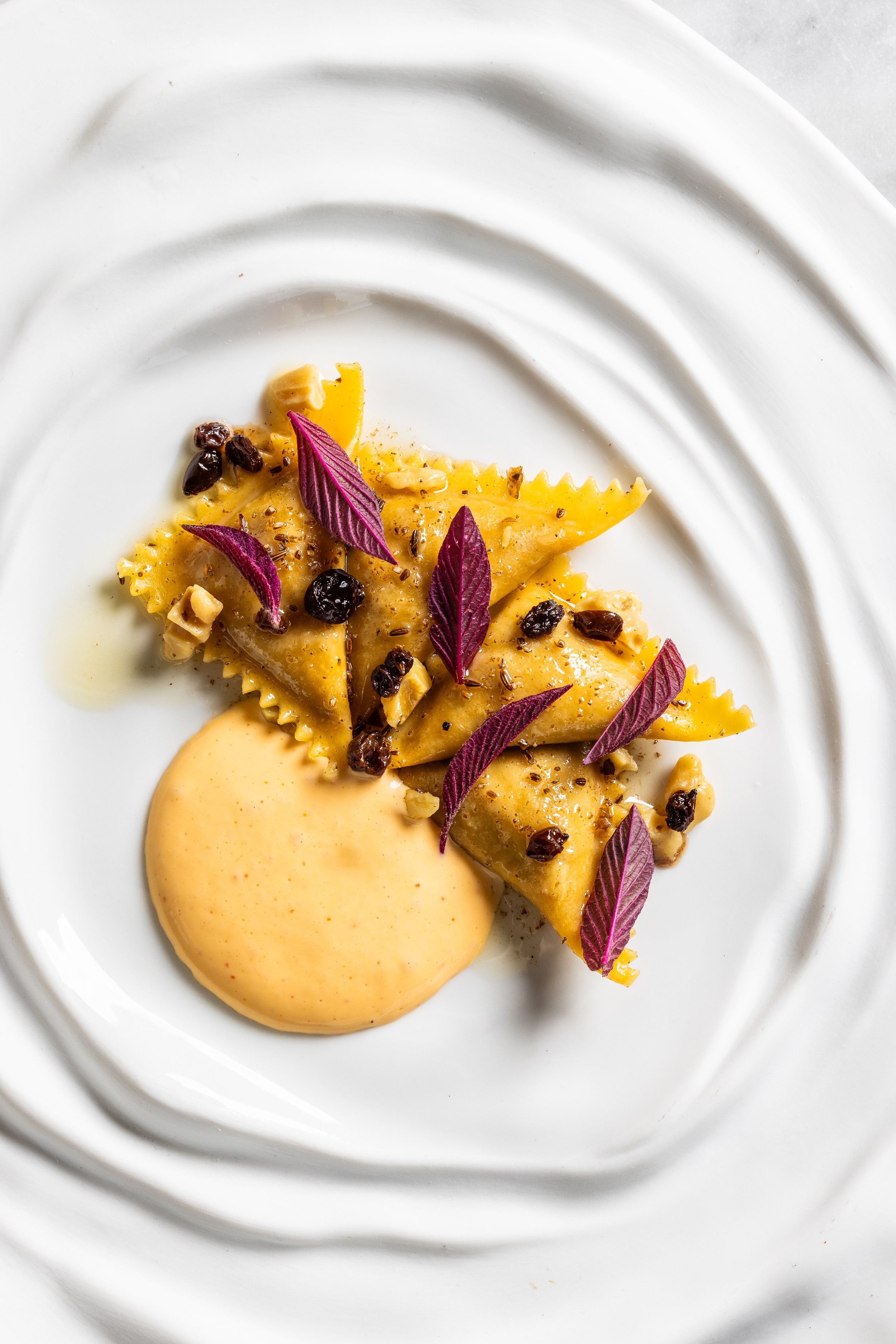 Plate of ravioli pasta garnished with purple leaves, black pepper, and nuts, with a side of creamy yellow sauce.
