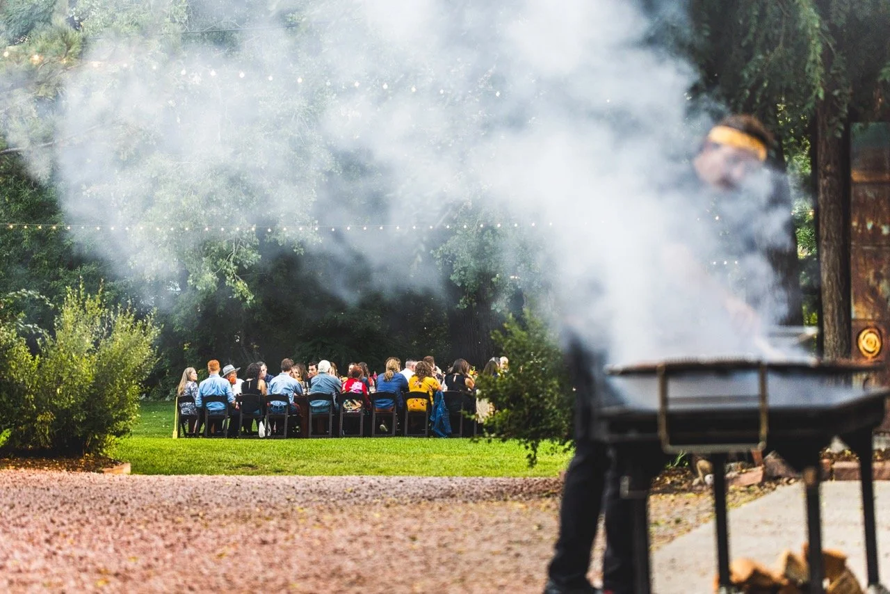People gathered outdoors around a table, with a person grilling food in the foreground and smoke rising from the grill.