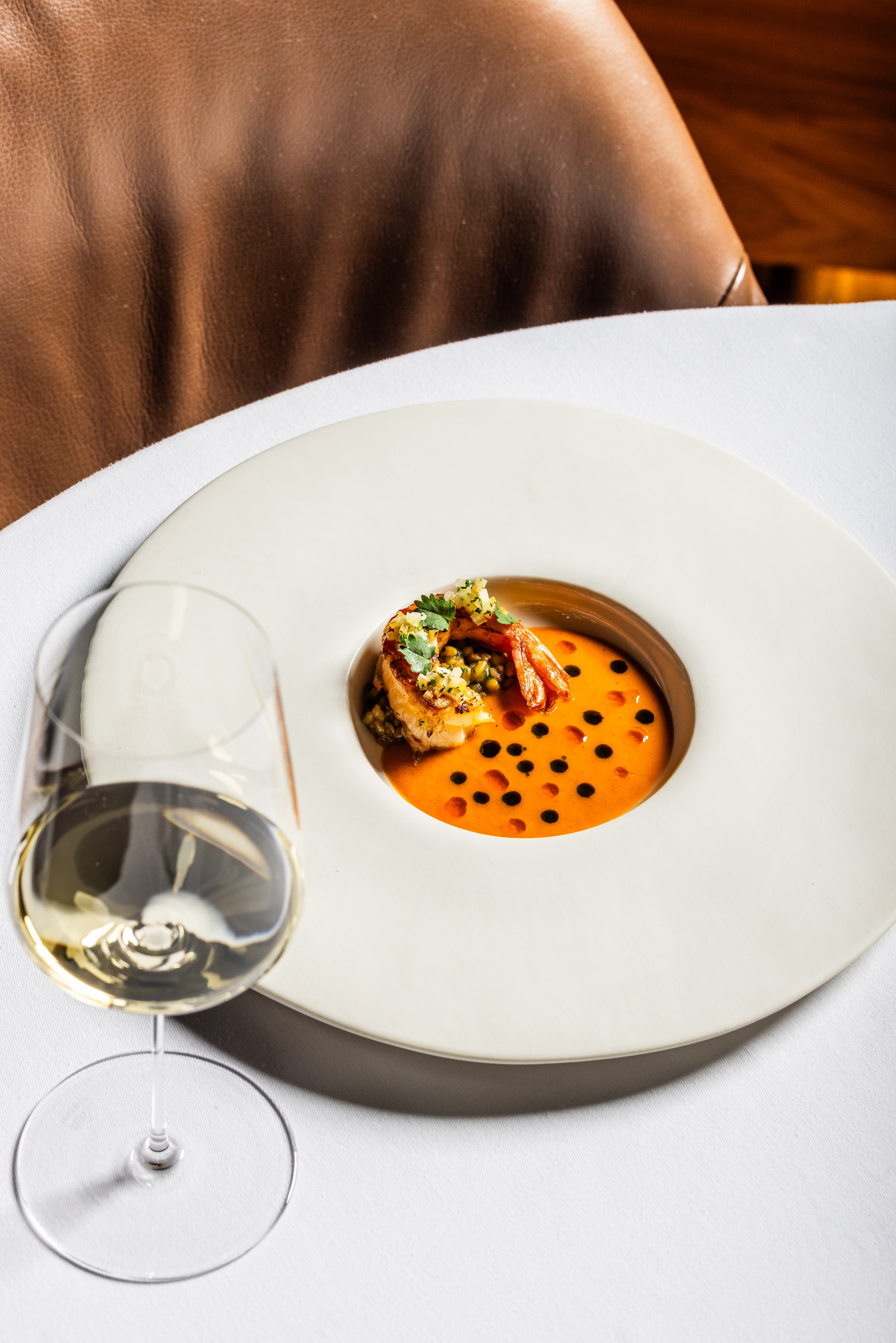 A gourmet dish with a shrimp, garnished with herbs and a pistachio topping, sits in an orange sauce with black dots on a white plate, accompanied by a glass of white wine.
