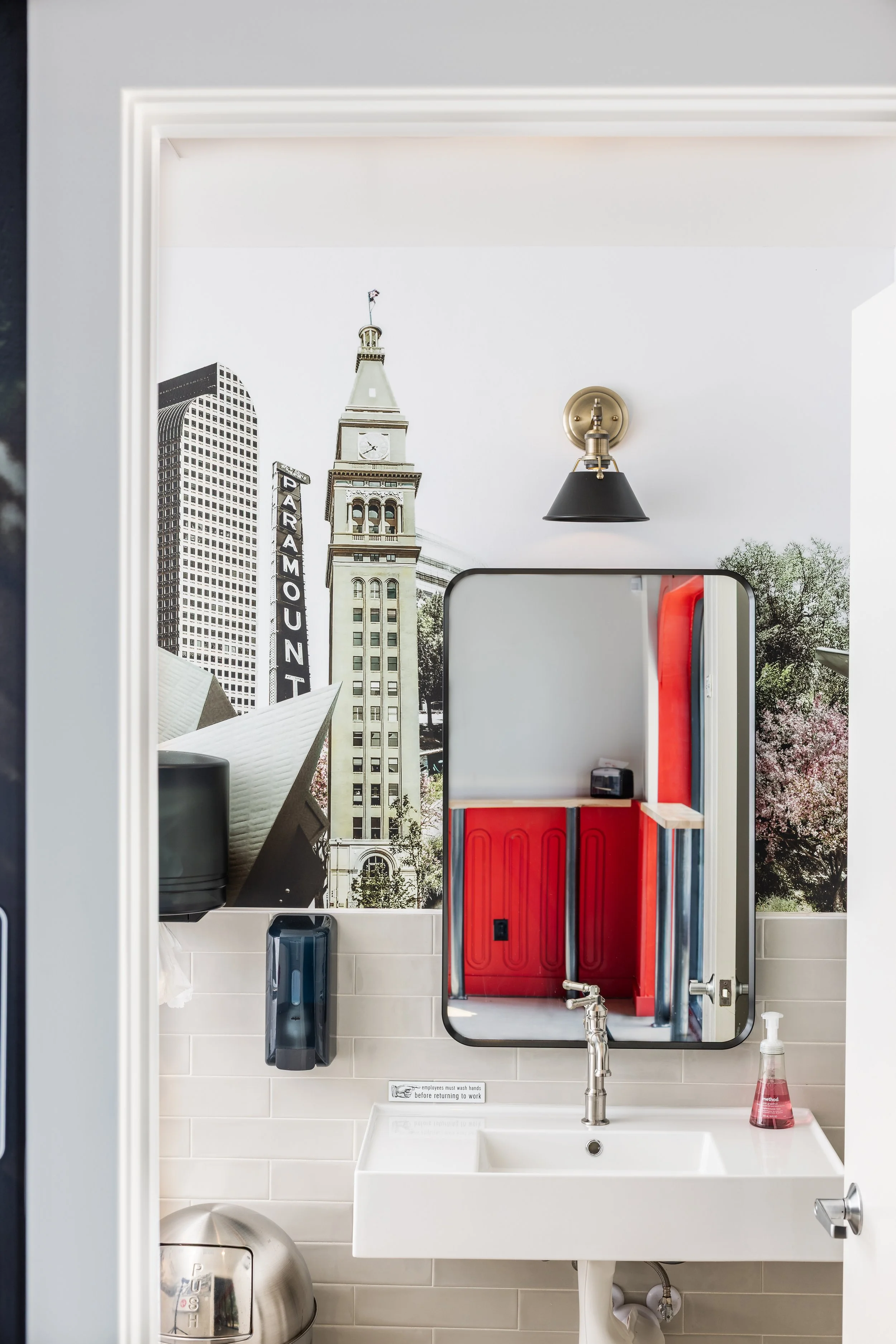 Restroom with a mirror, sink, soap dispenser, and wall decor featuring a cityscape mural, including a tall clock tower and modern buildings.