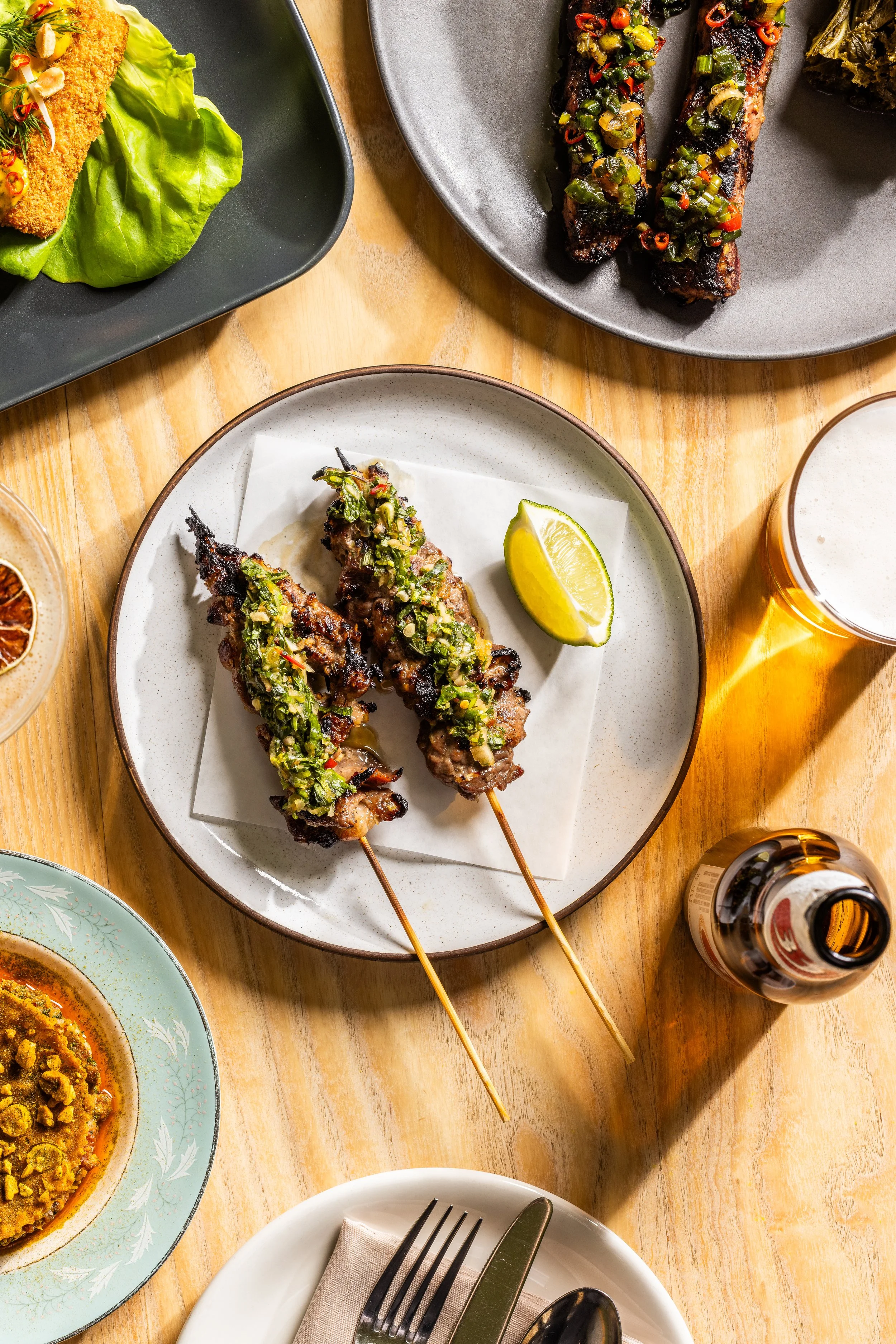 Two skewers of grilled meat topped with green chopped herbs and diced vegetables, served with lime wedges on a white plate.