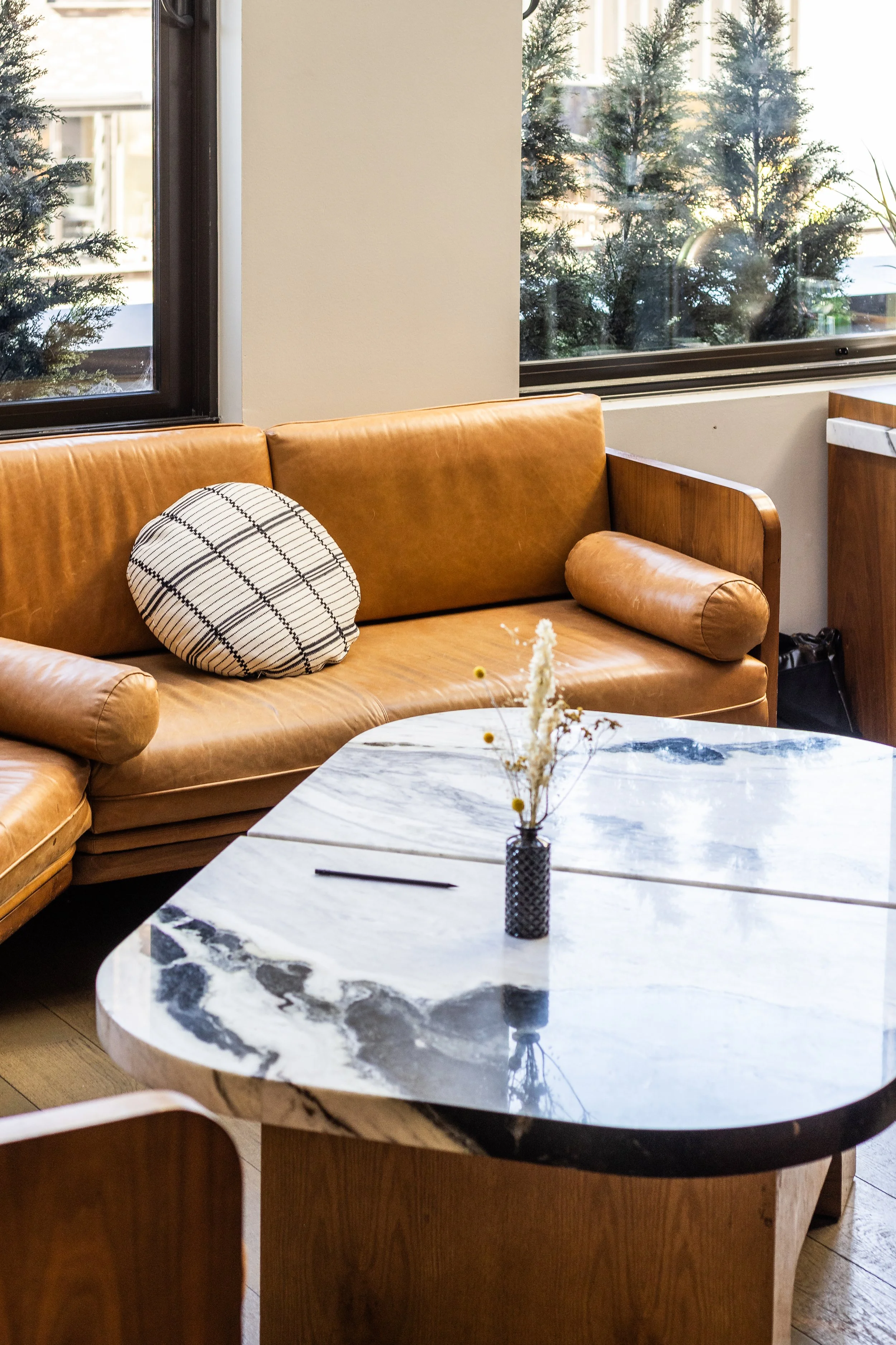 A cozy indoor seating area with a bronze leather couch, a decorative round pillow, a cylindrical armrest, a marble-topped table with a small vase of dried flowers, and windows showing outdoor trees.