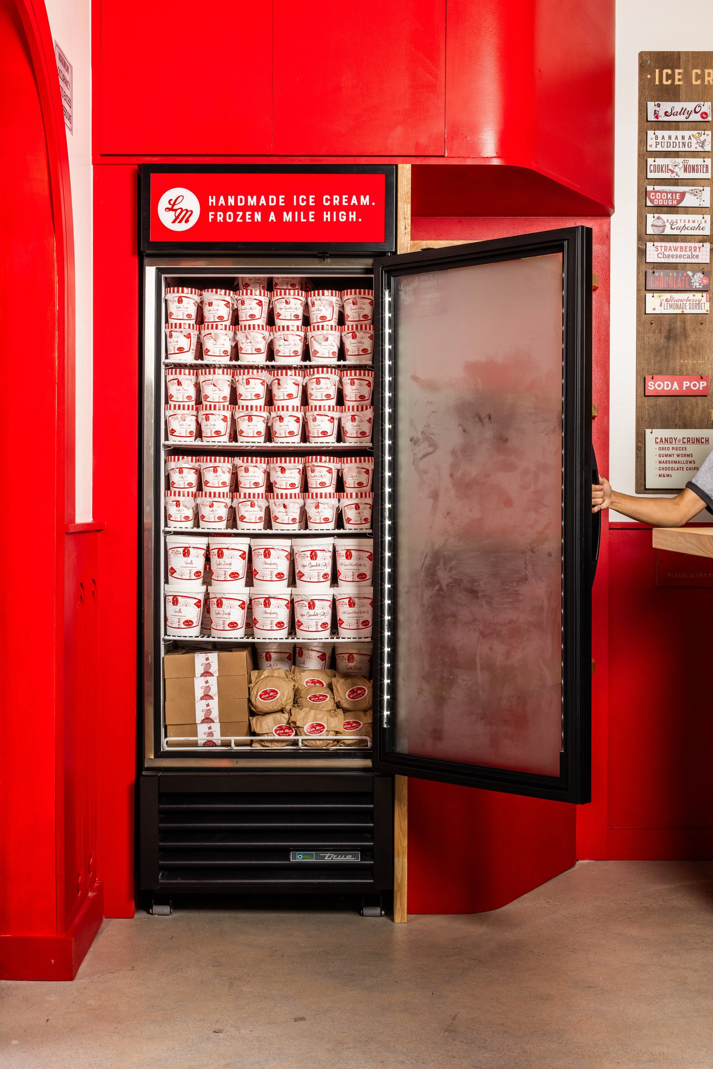 A commercial freezer filled with packaged ice cream containers, with a sign above that reads 'Handmade Ice Cream. Frozen a Mile High.' on a red wall.