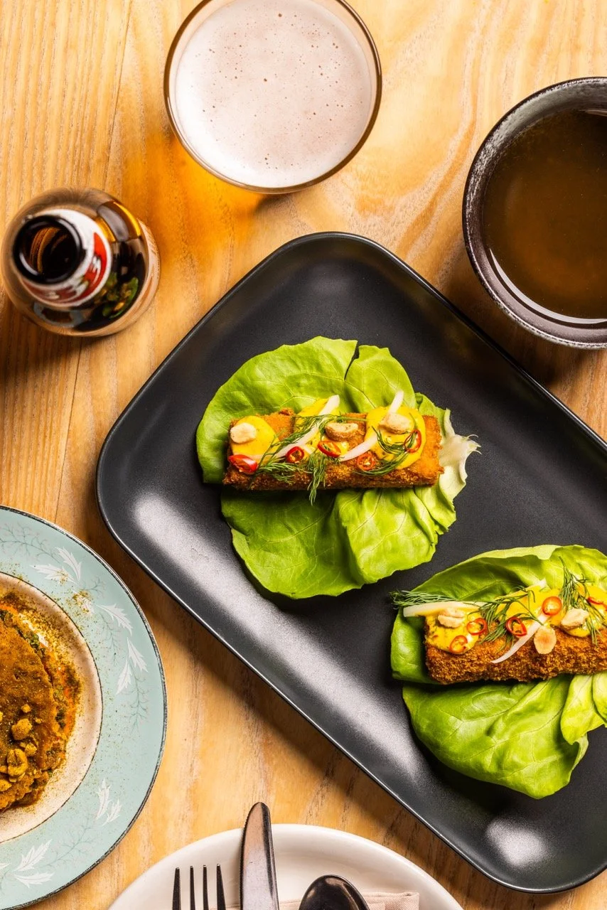 A black plate with two lettuce wraps filled with fried fish, chopped red chili, garlic slices, and green herbs on a wooden table. Surrounding the plate are a glass of beer, a bowl of soup, a beer bottle, and a partially visible plate of food.