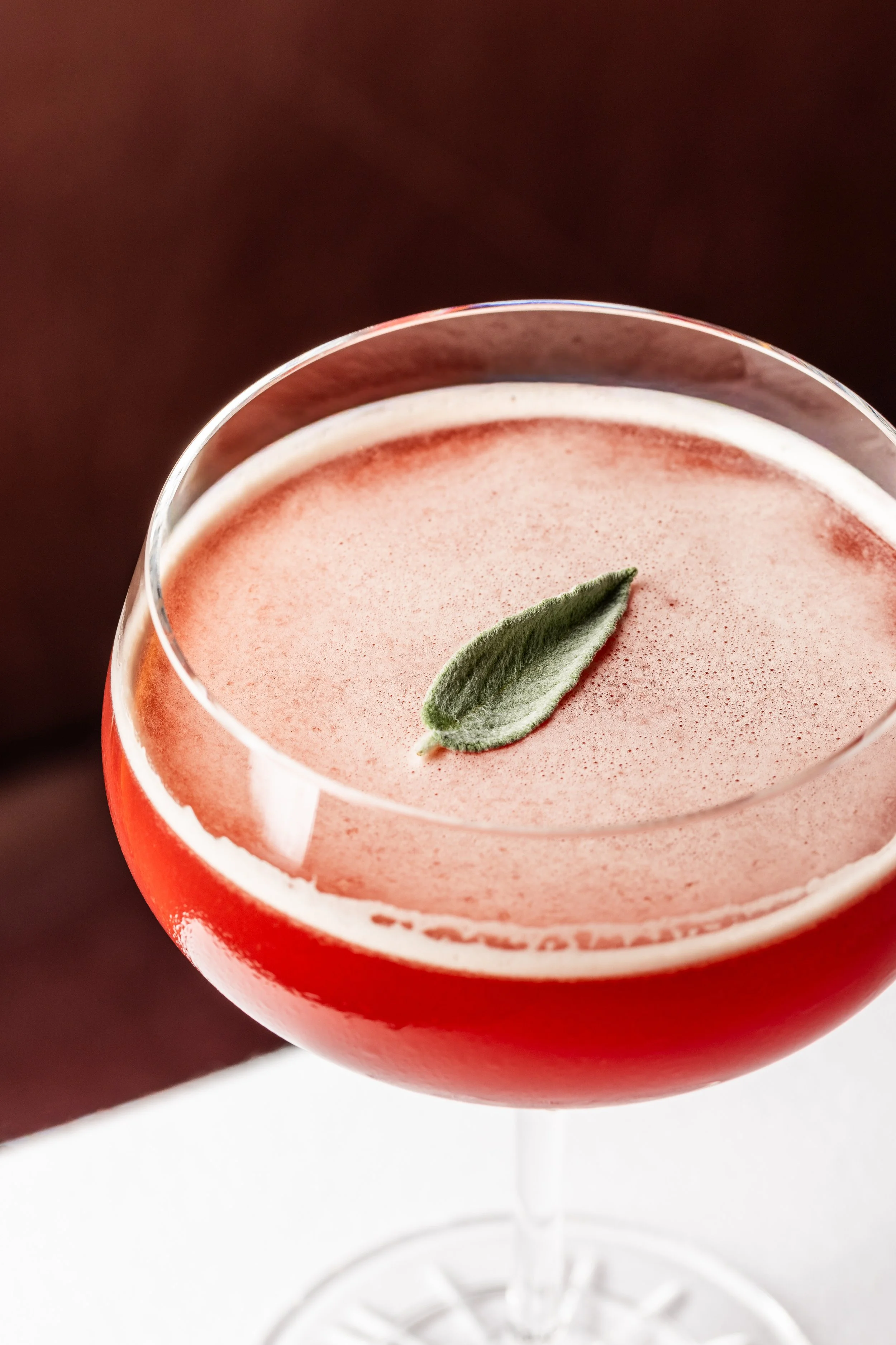 A red cocktail in a stemmed glass garnished with a sage leaf.