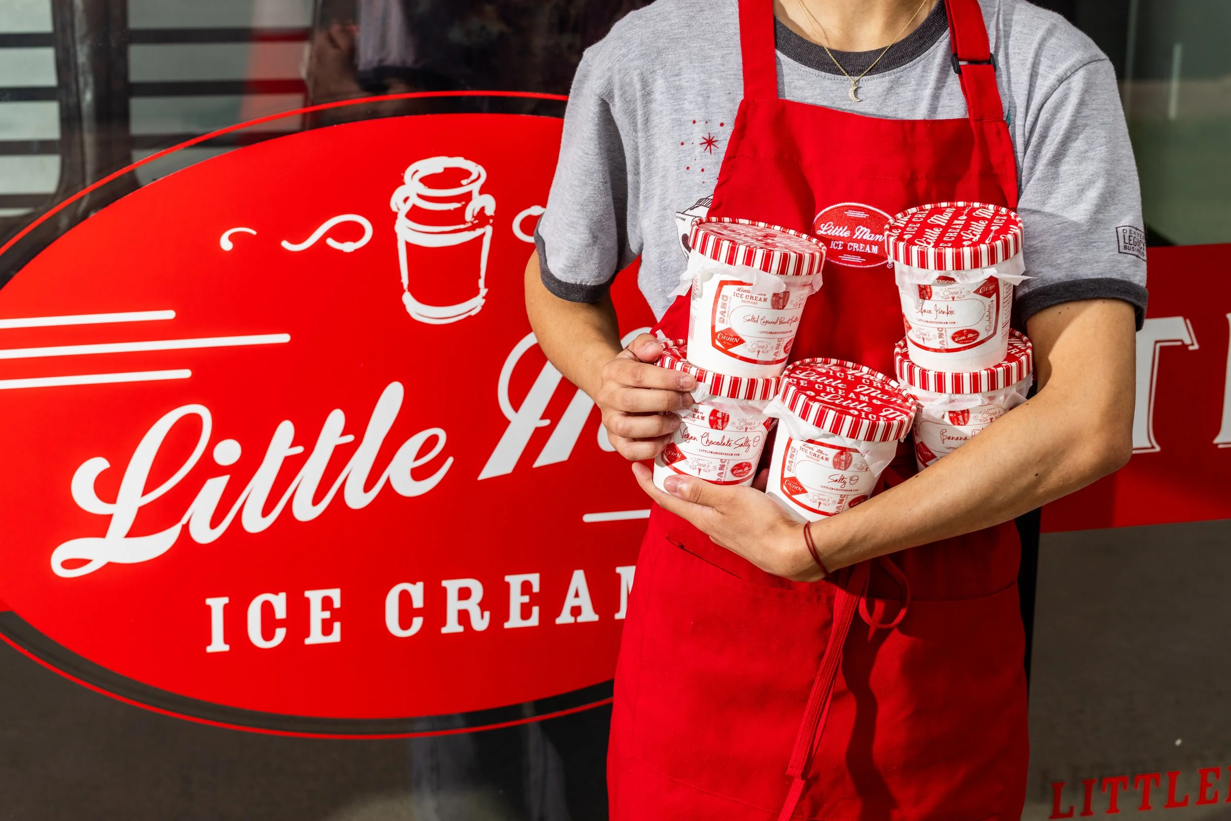 Person wearing a gray t-shirt and red apron holding several containers of Little Man Ice Cream in front of the Little Man Ice Cream store logo.