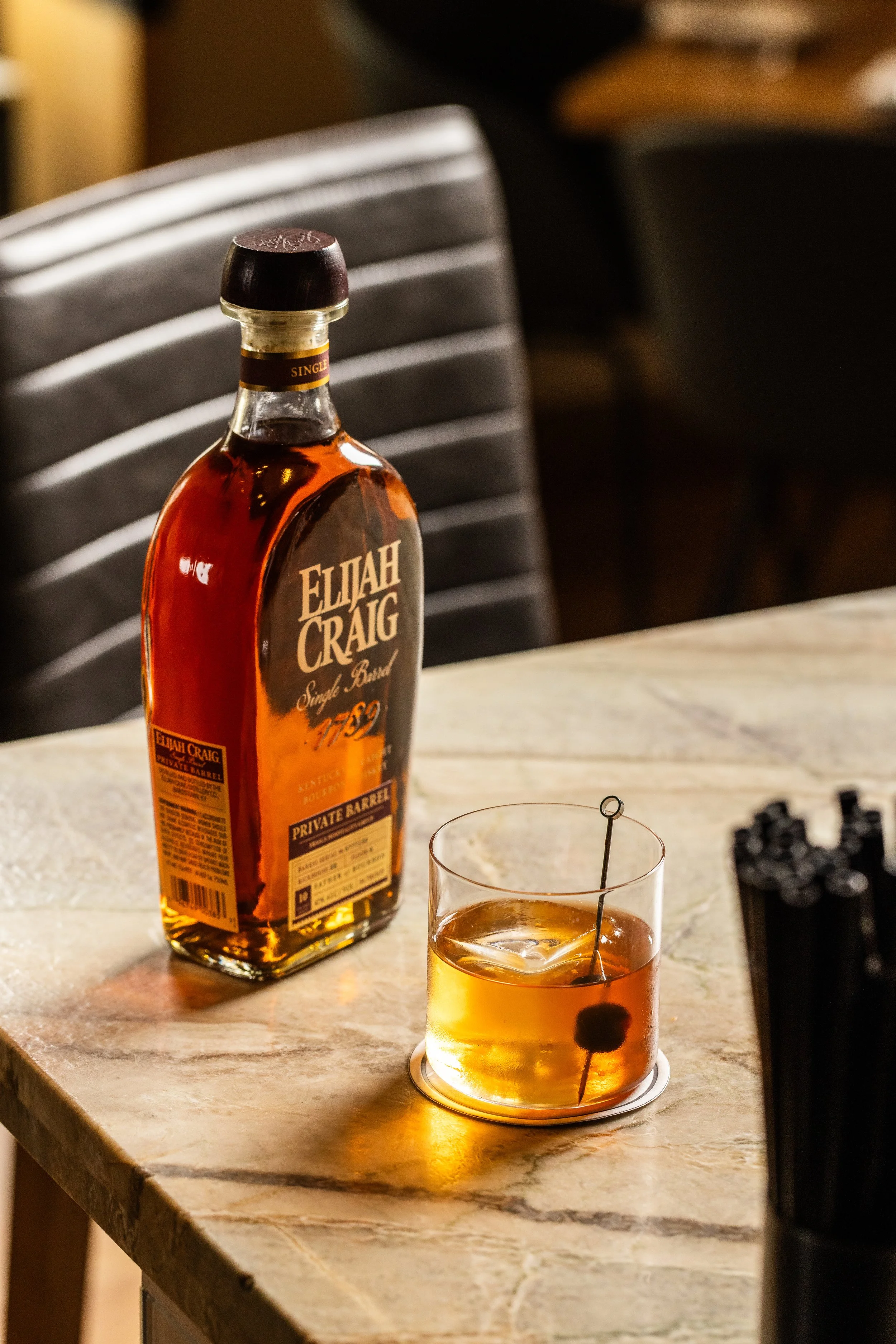 A bottle of Elijah Craig bourbon whiskey and a glass with whiskey and a cherry, garnished with a cocktail pick, on a marble countertop.
